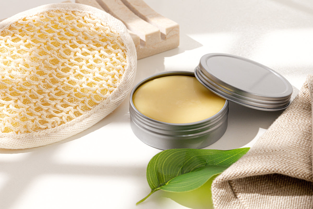 Open silver tin filled with pale yellow balm, surrounded by a cream-colored exfoliating bath sponge, handmade soap bars, a green leaf, and beige linen fabric on a sunlit white surface.