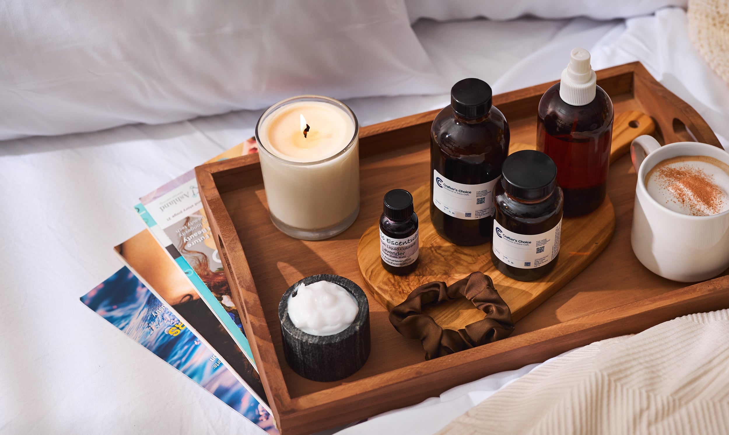 Cozy bedroom tray with a lit candle, skincare bottles, cream jar, silk scrunchie, and a latte on a wooden tray beside magazines on a bed.