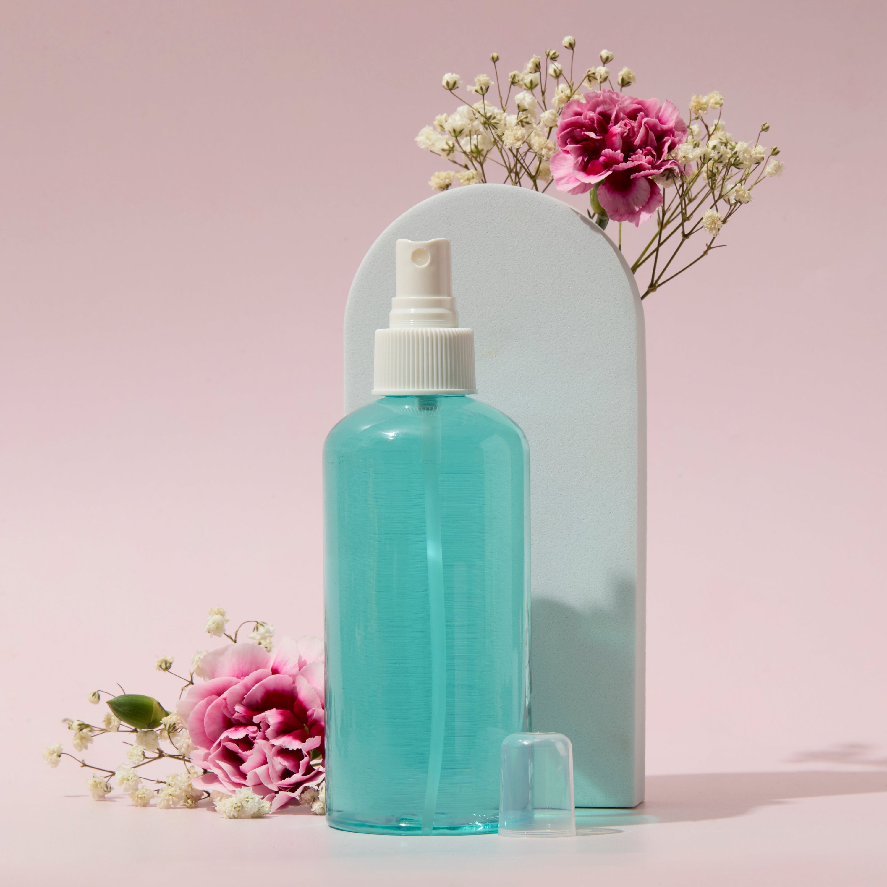 Blue liquid spray in clear bottle with white sprayer cap displayed with pink flowers on soft pastel background