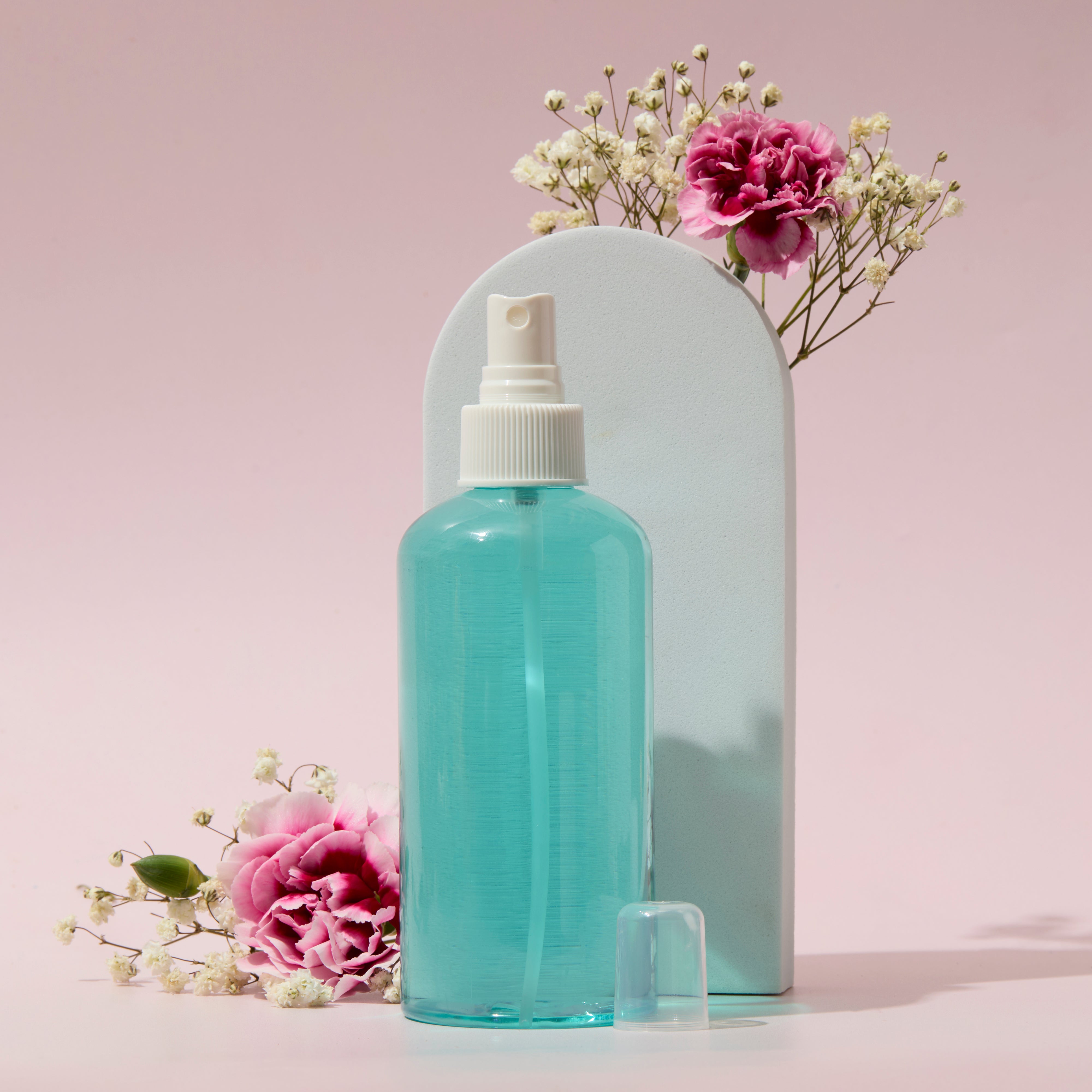 Blue liquid spray in clear bottle with white sprayer cap displayed with pink flowers on soft pastel background