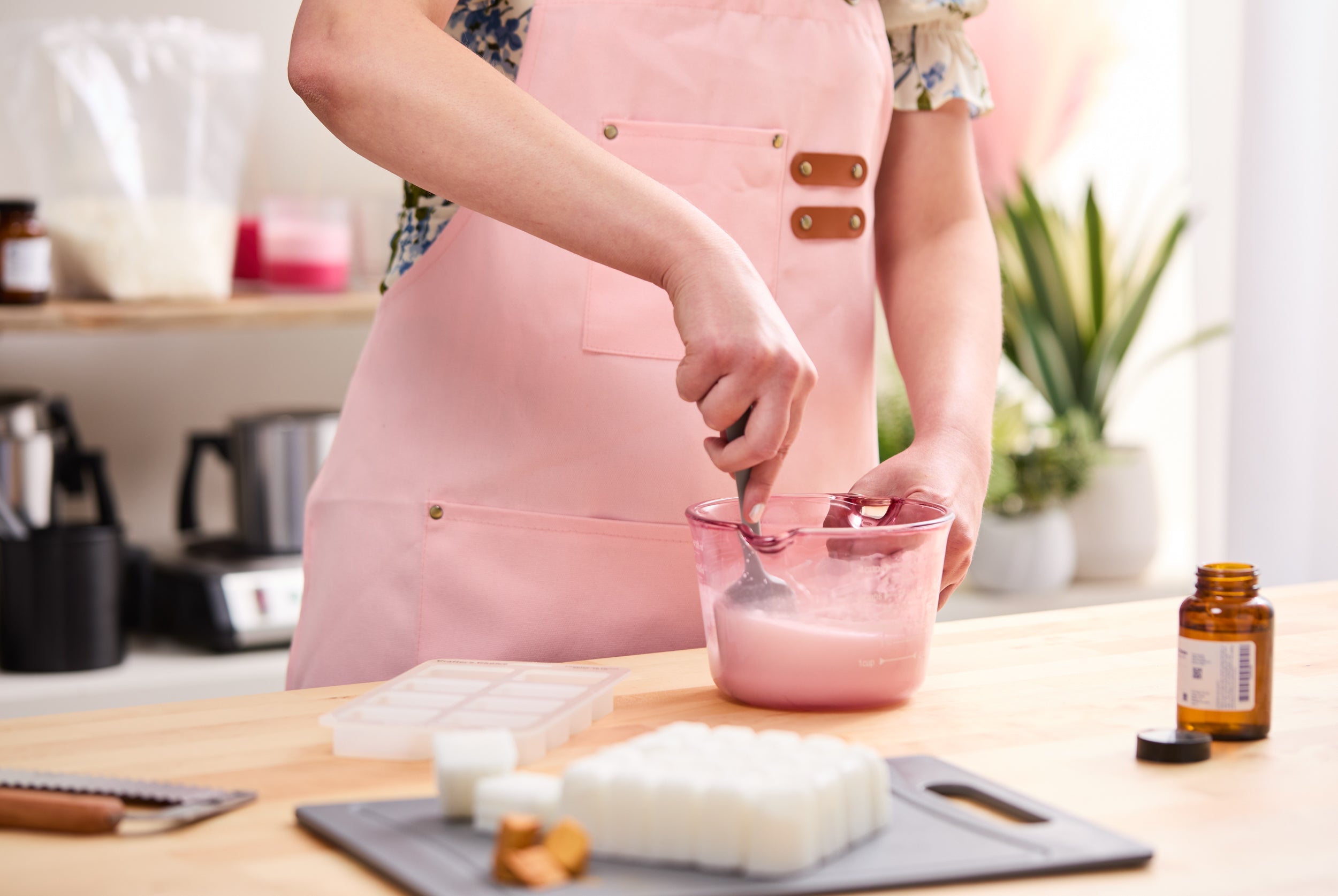 Person wearing a pink apron stirring melted wax in a pink measuring cup at a crafting table, with soap or wax melts, a mold tray, and fragrance oil nearby in a bright home workspace.