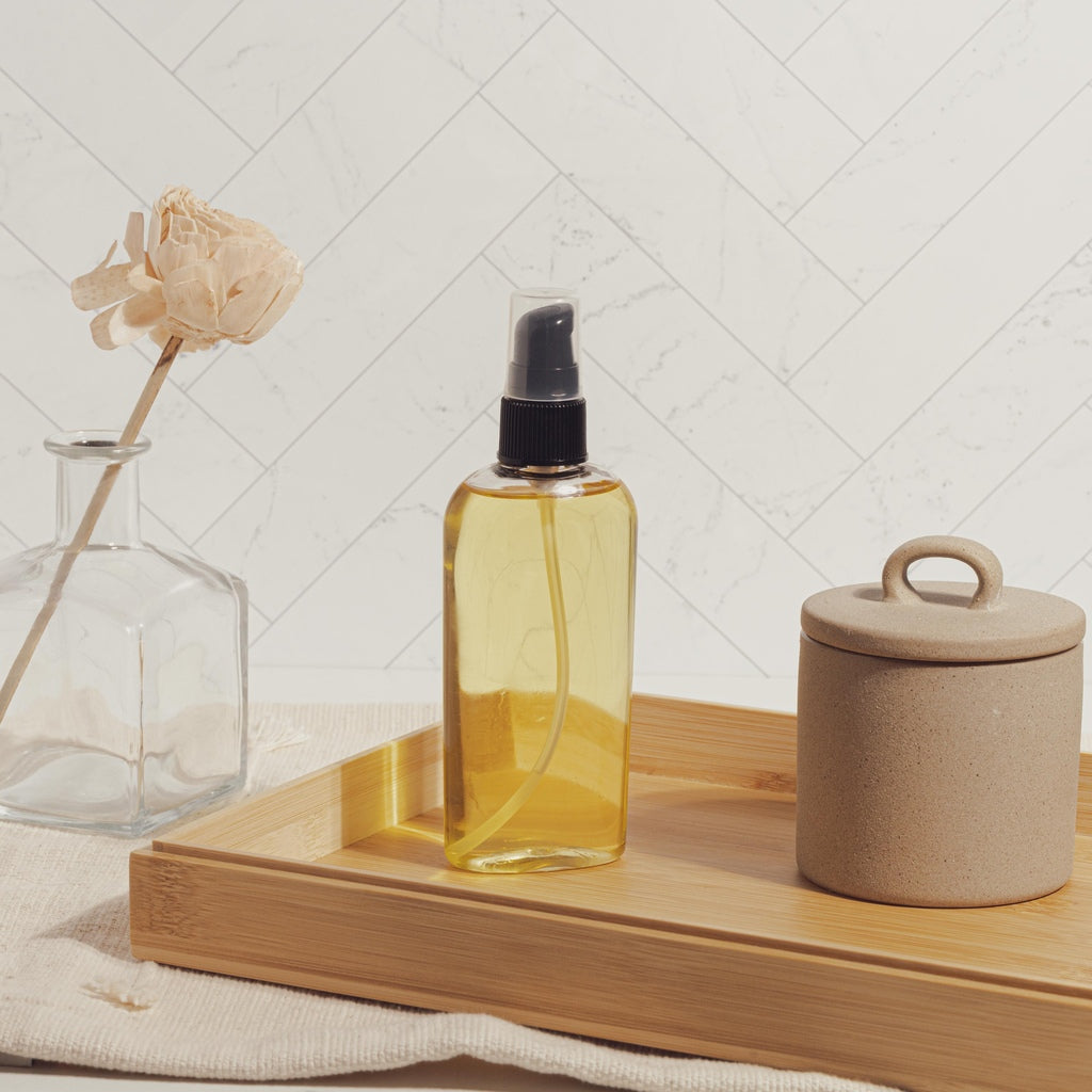 Golden body oil in clear spray bottle on wooden tray with ceramic jar and glass vase in neutral bathroom setting