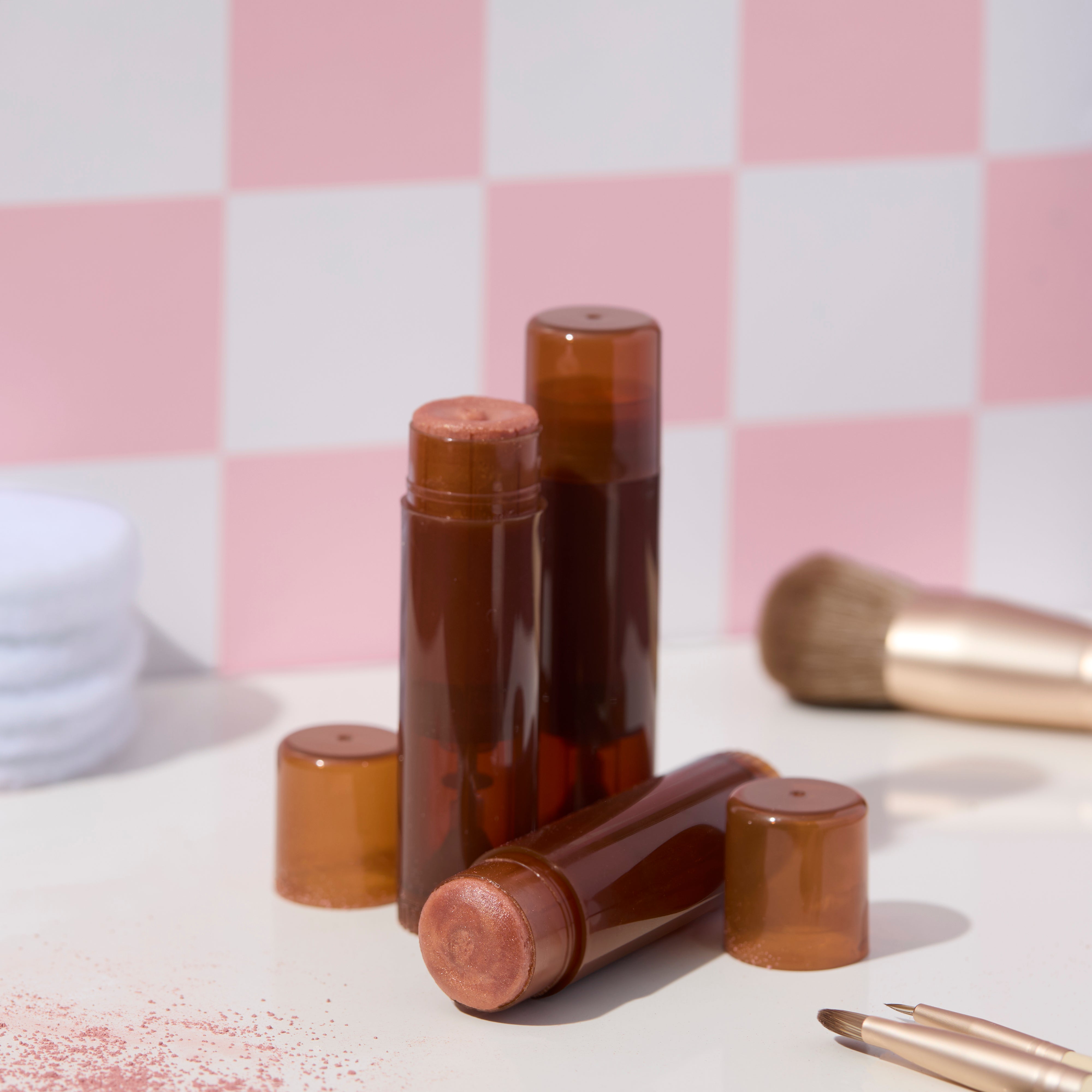 Amber twist up cosmetic tubes with bronze shimmer balm displayed on white surface with pink checkered background