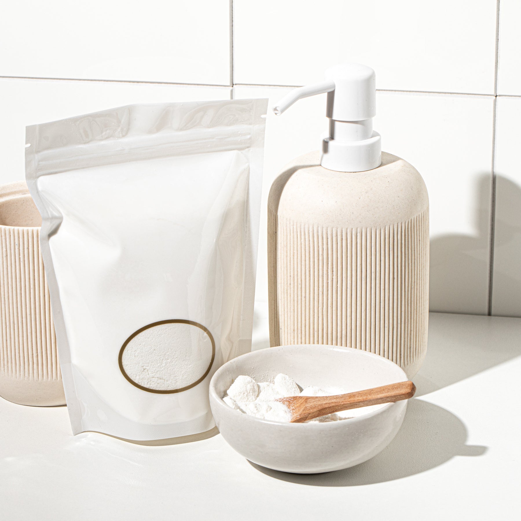 White laundry detergent powder in bowl with wooden scoop beside refill pouch and pump bottle on bathroom counter
