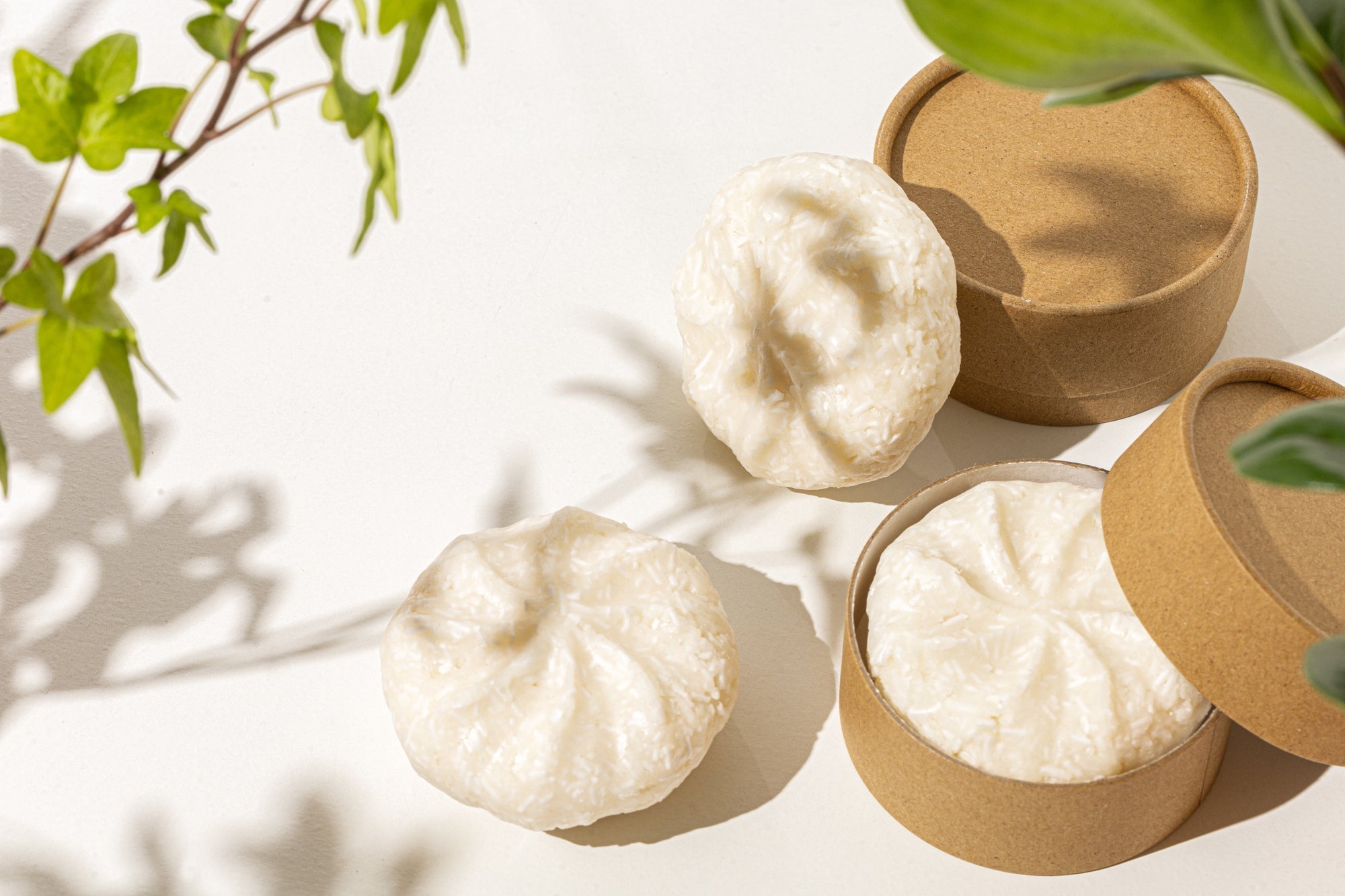 Minimal flat lay of creamy white shampoo bars with textured tops, styled on a light surface alongside kraft paper round containers and soft green leaves casting natural shadows.
