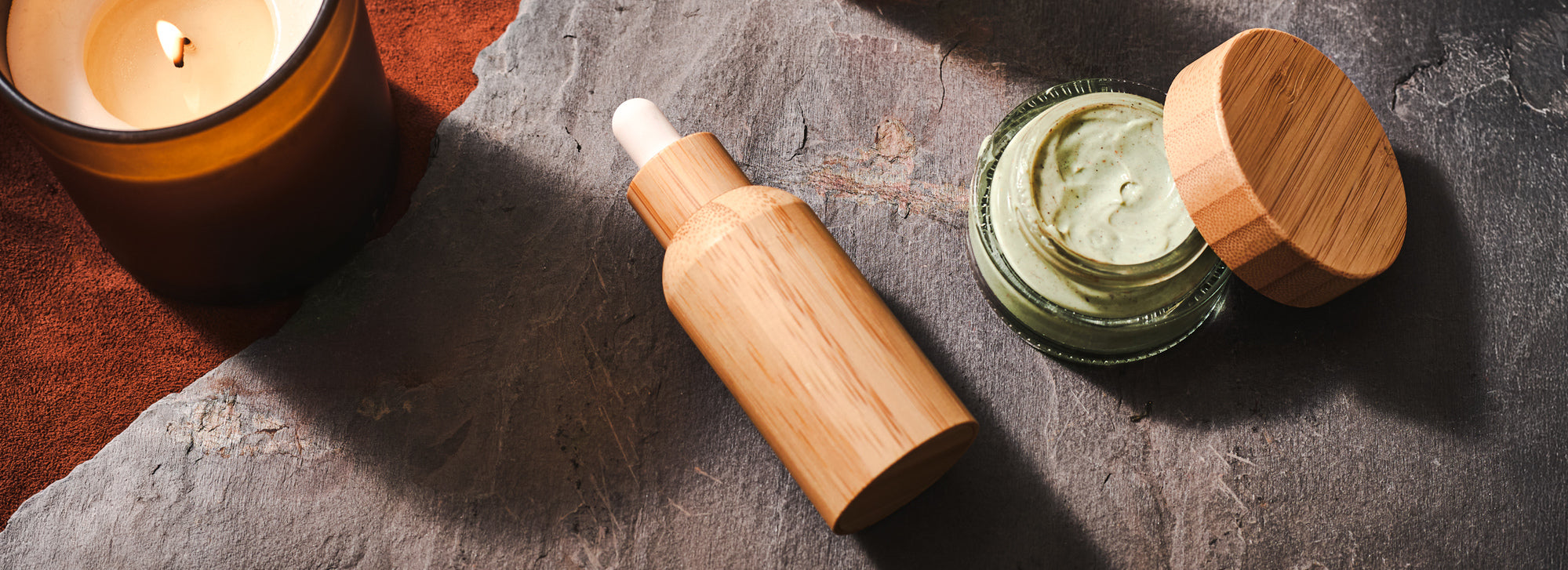 Bamboo skincare dropper bottle beside an open jar of green cream and a lit candle on a textured stone surface.