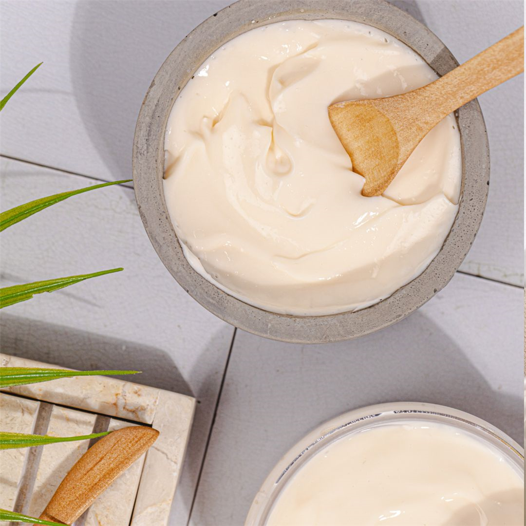 Creamy body lotion in open jar and bowl with wooden spatula on tiled surface with plant leaf and soap dish