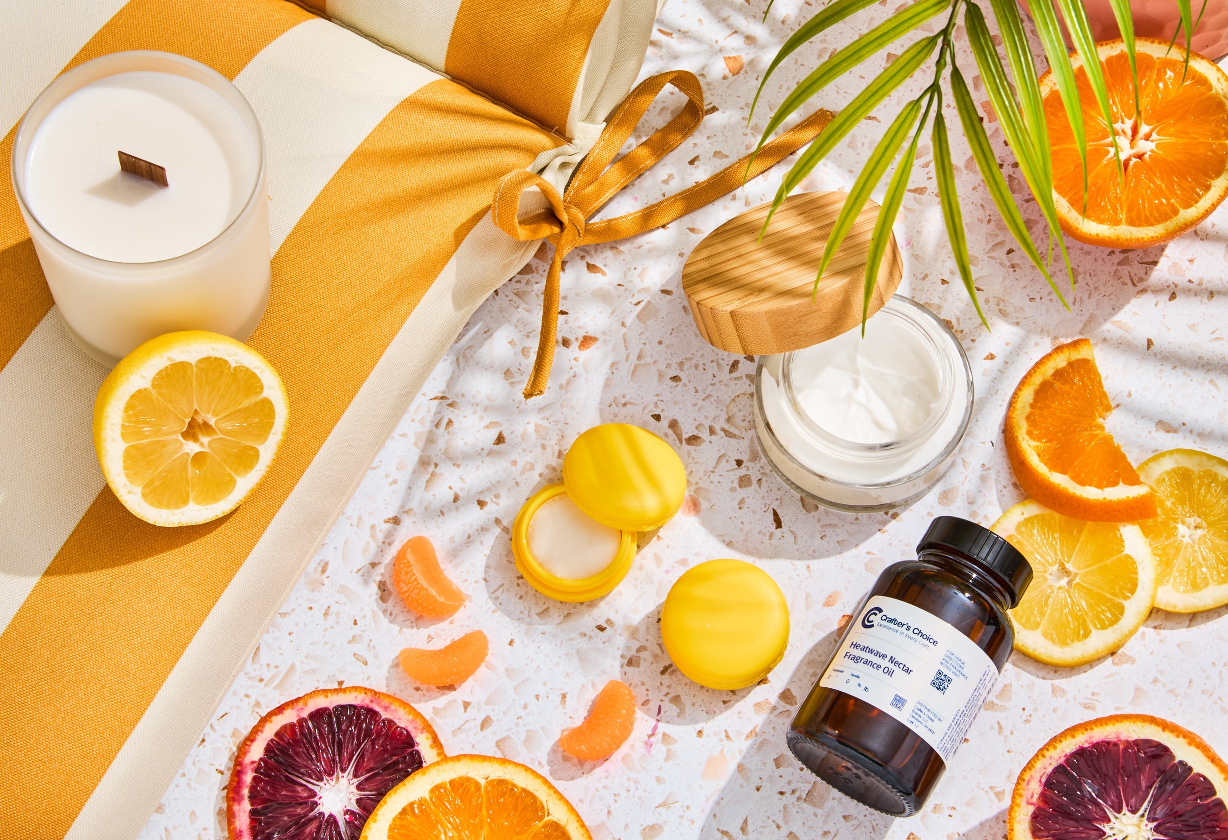 Bright citrus-themed flat lay featuring a white candle, jar of body cream, yellow lip balm containers, and a fragrance oil bottle surrounded by sliced lemons, oranges, blood oranges, and citrus segments on a terrazzo surface.