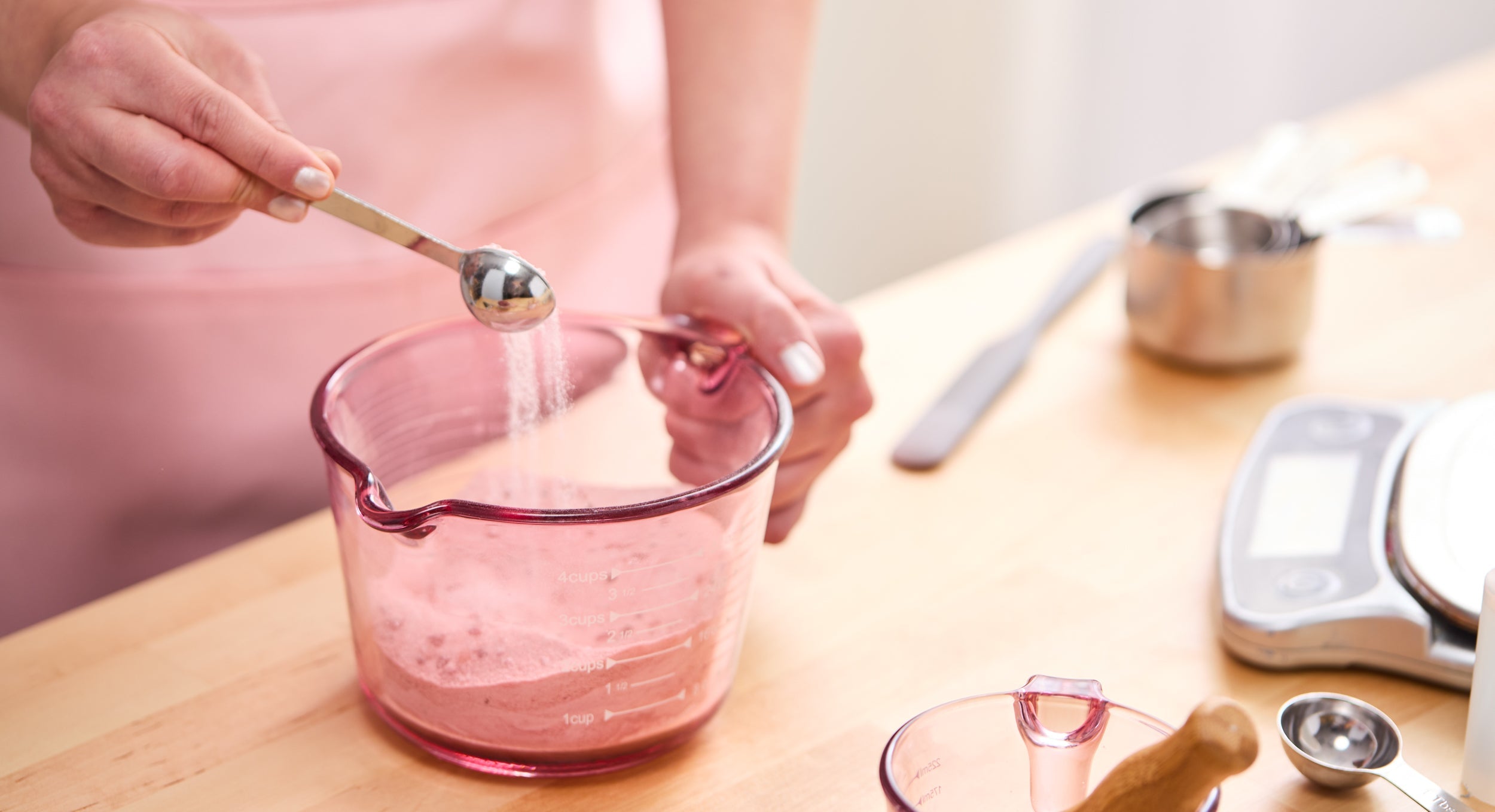Close-up of a person in a pink apron adding a spoonful of fine powder into a pink measuring cup filled with a dry bath mix, with measuring tools and a scale on the wooden workspace.