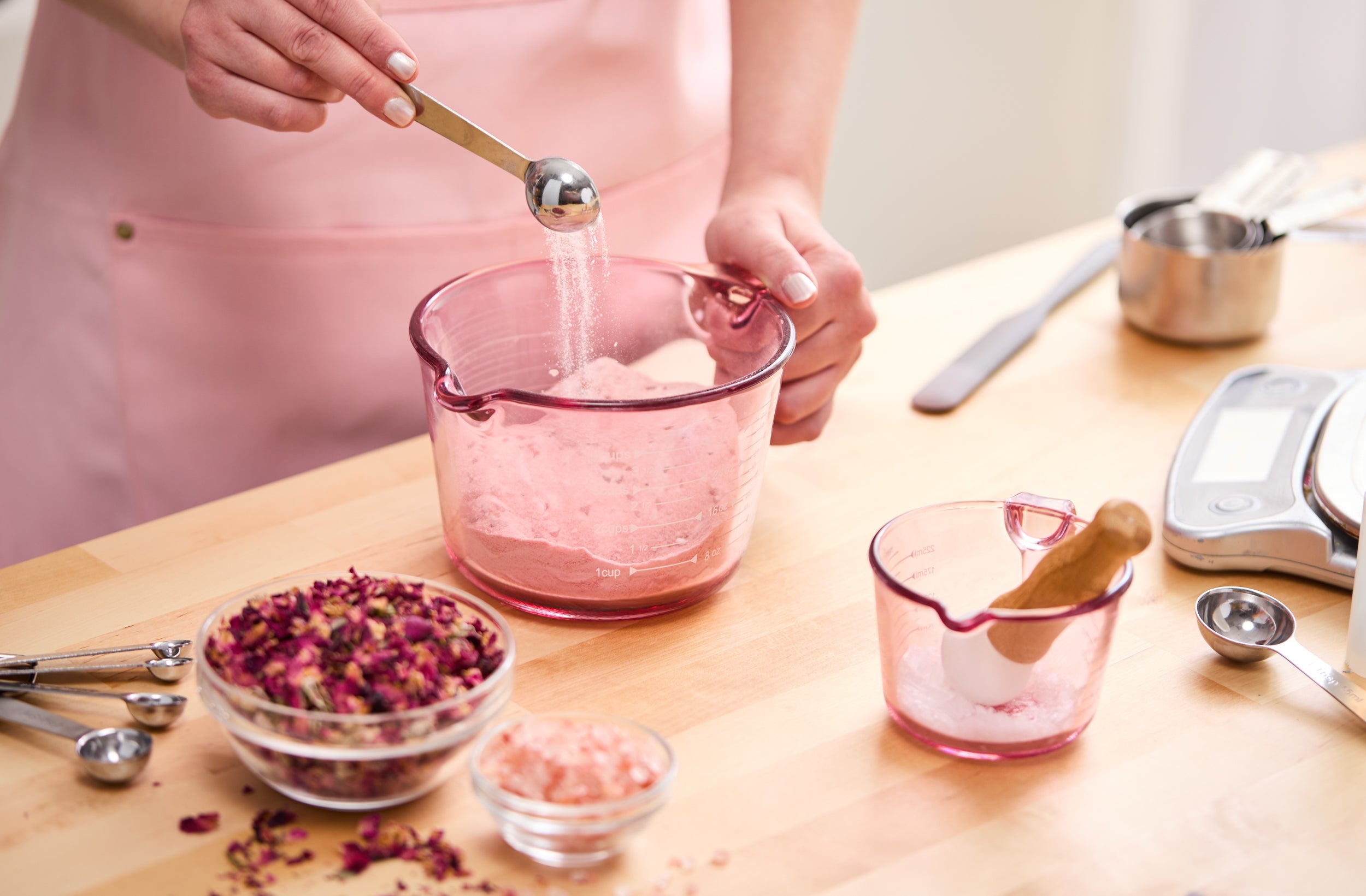 Person in a pink apron adding powdered ingredients to a pink measuring cup, surrounded by bowls of dried rose petals and pink salts, measuring spoons, and a small mortar and pestle on the workspace.