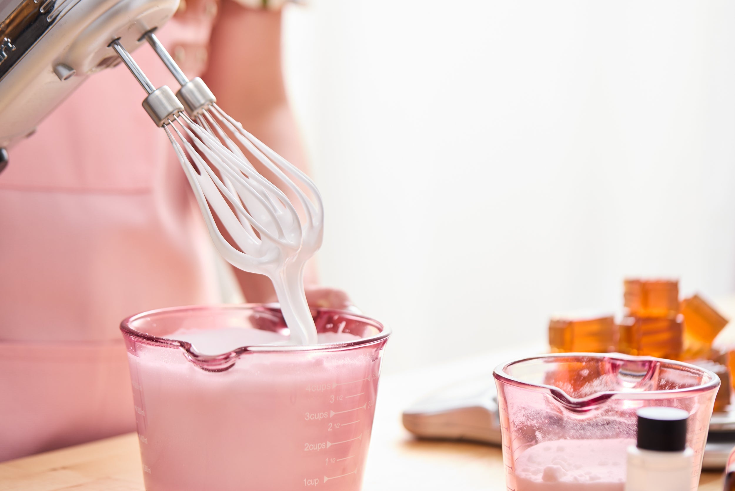 Close-up of a hand mixer lifting thick, whipped white body-care mixture from a pink measuring cup, with additional ingredients and melted soap blocks on the crafting table in a bright workspace.