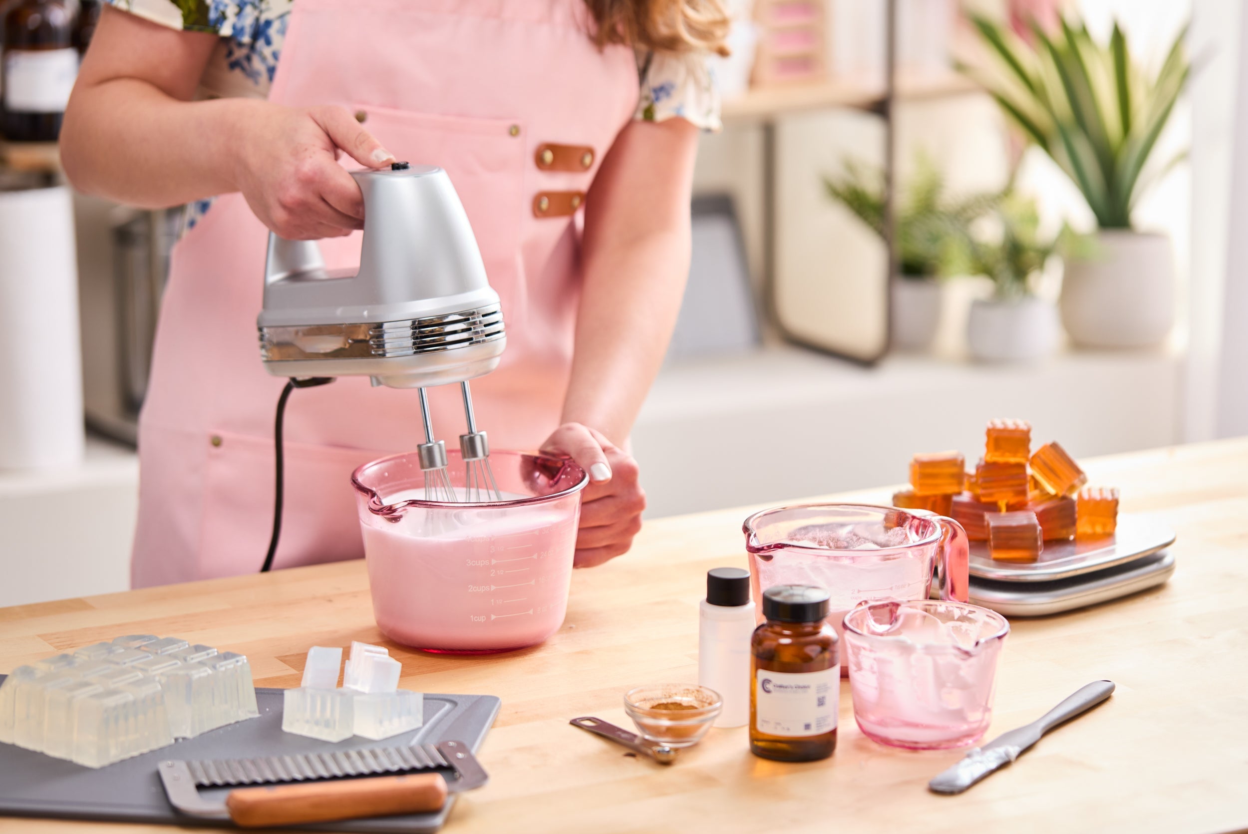 Person in a pink apron using a hand mixer to blend a pink soap or body-care mixture in a glass measuring cup, surrounded by melt-and-pour soap blocks, fragrance oils, colorants, and crafting tools on a bright workspace table.