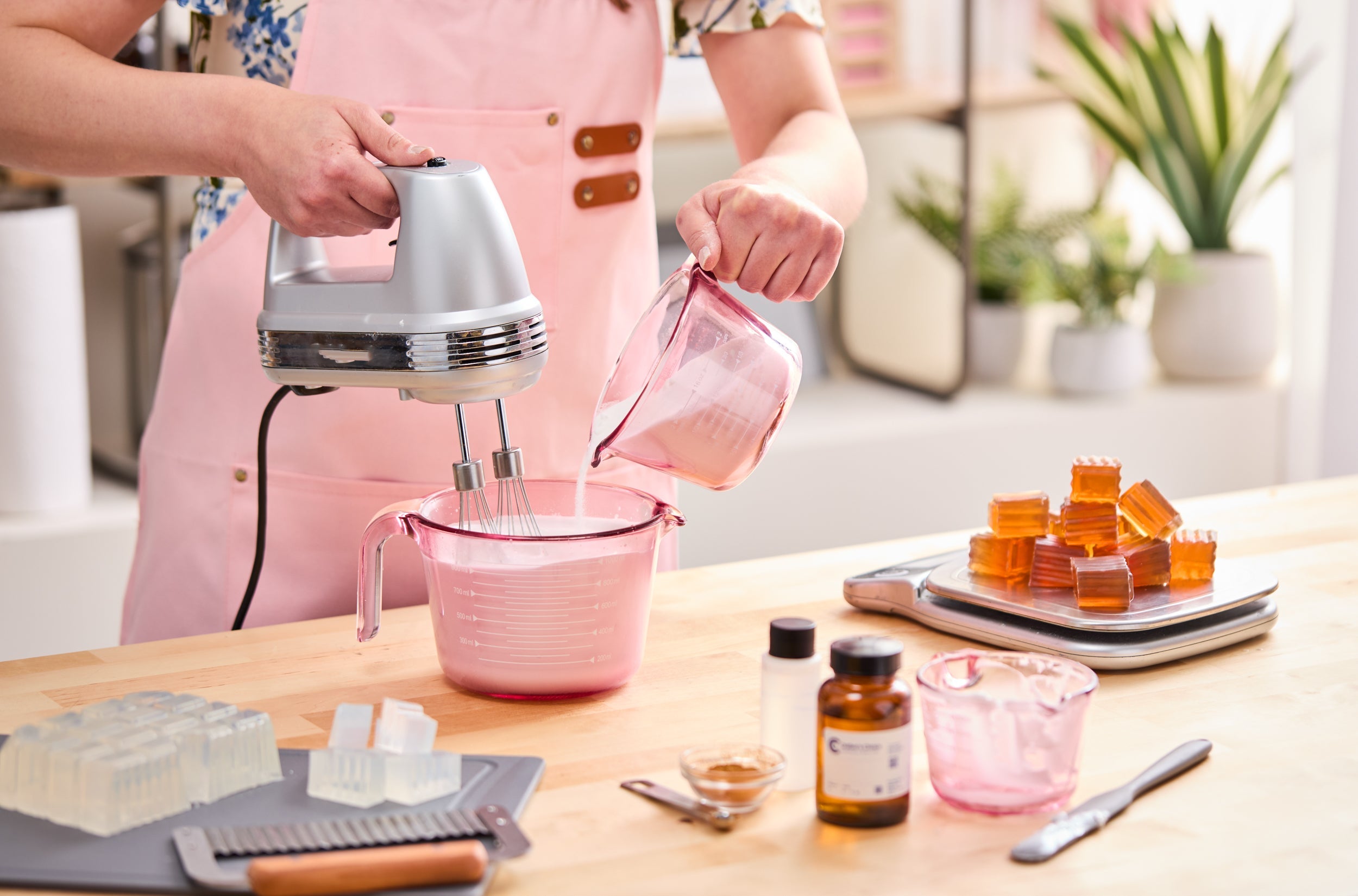 Person in a pink apron using a hand mixer while pouring a white mixture into a larger pink measuring cup, surrounded by soap-making supplies including clear and amber soap cubes, amber bottles, and measuring tools.