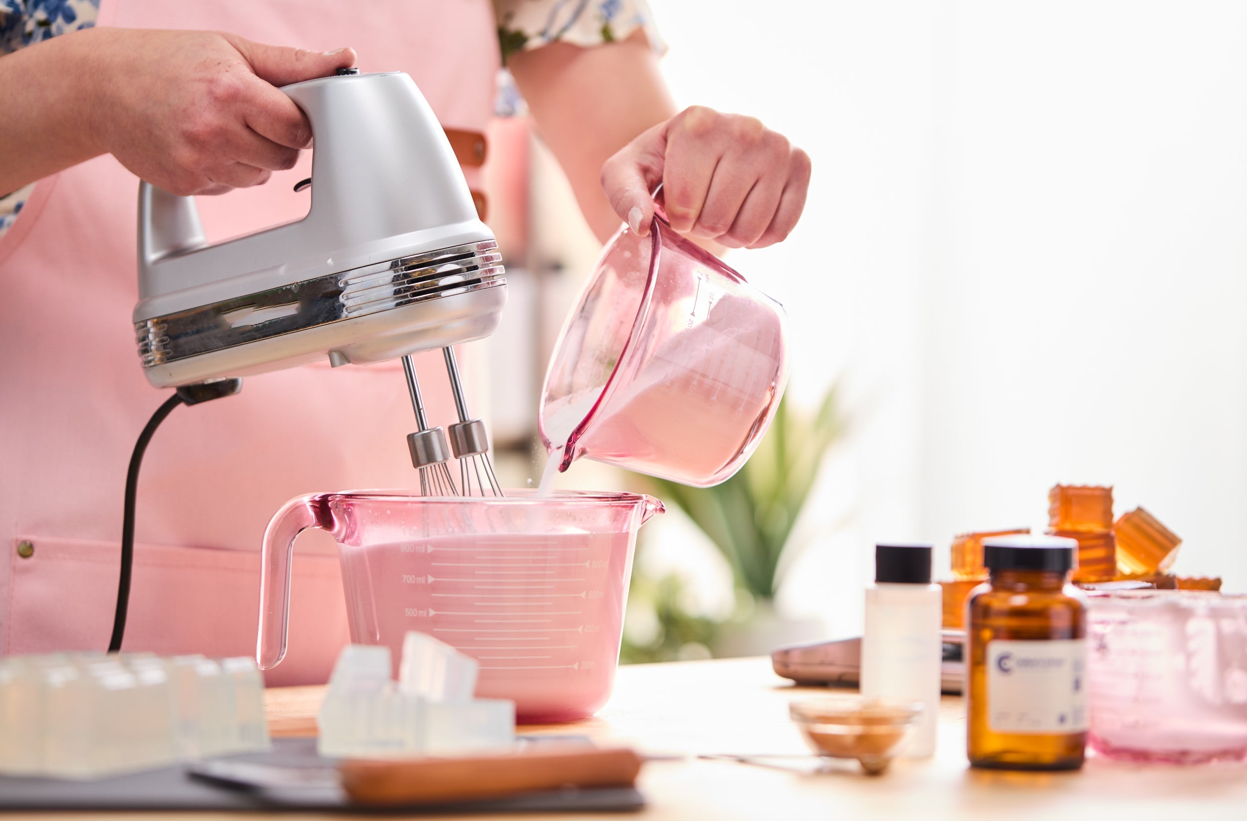 Person in a pink apron using a hand mixer while pouring a creamy pink mixture into a larger pink measuring cup, surrounded by soap-making supplies, amber bottles, and clear soap cubes on the table.