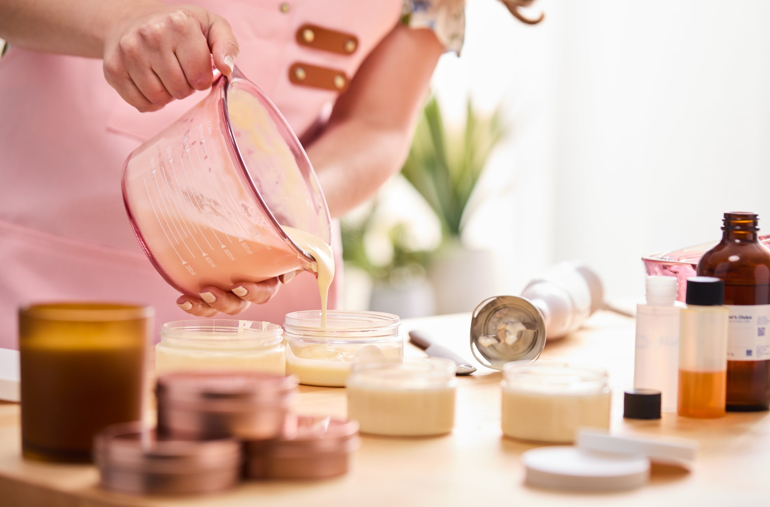 Person in a pink apron pouring creamy lotion mixture from a pink measuring cup into clear jars, surrounded by filled jars, lids, an immersion blender, and bottles of crafting ingredients on the table.