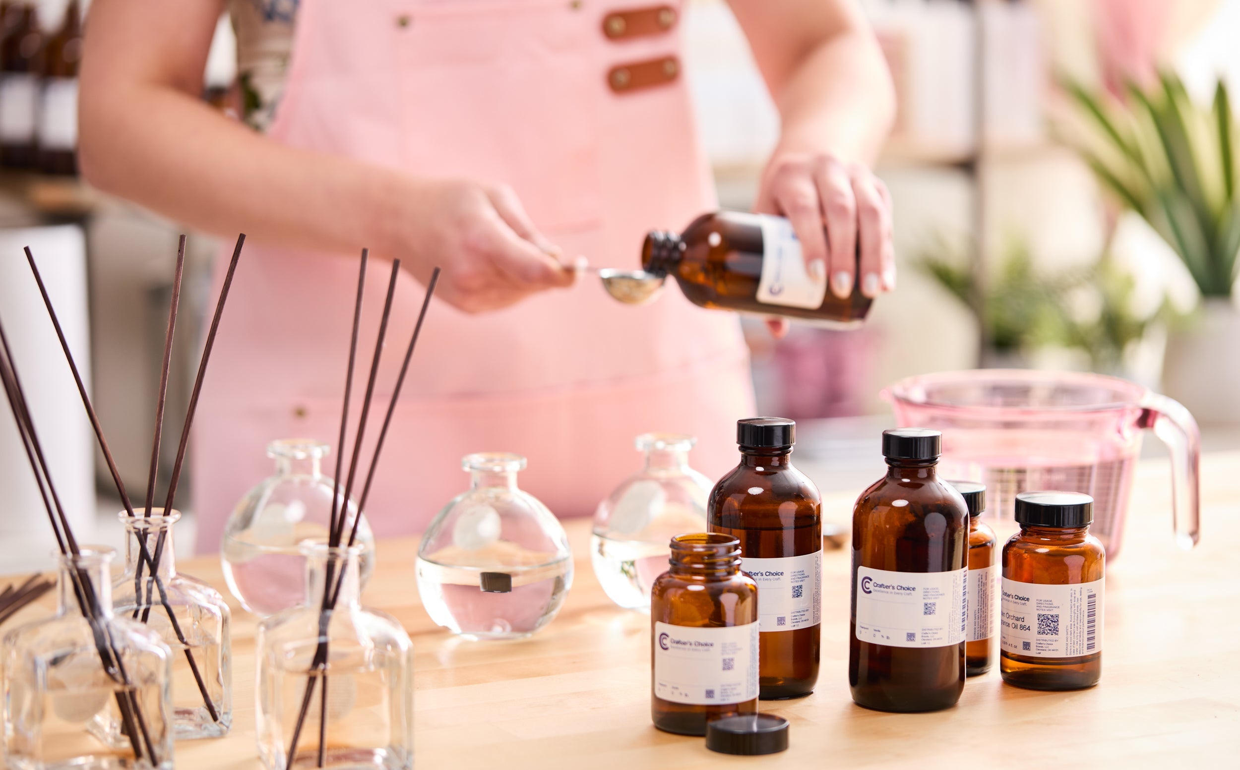 “Person in a pink apron measuring fragrance oil from an amber bottle into a metal spoon, with reed diffuser bottles filled with liquid and additional amber fragrance bottles arranged on the table.