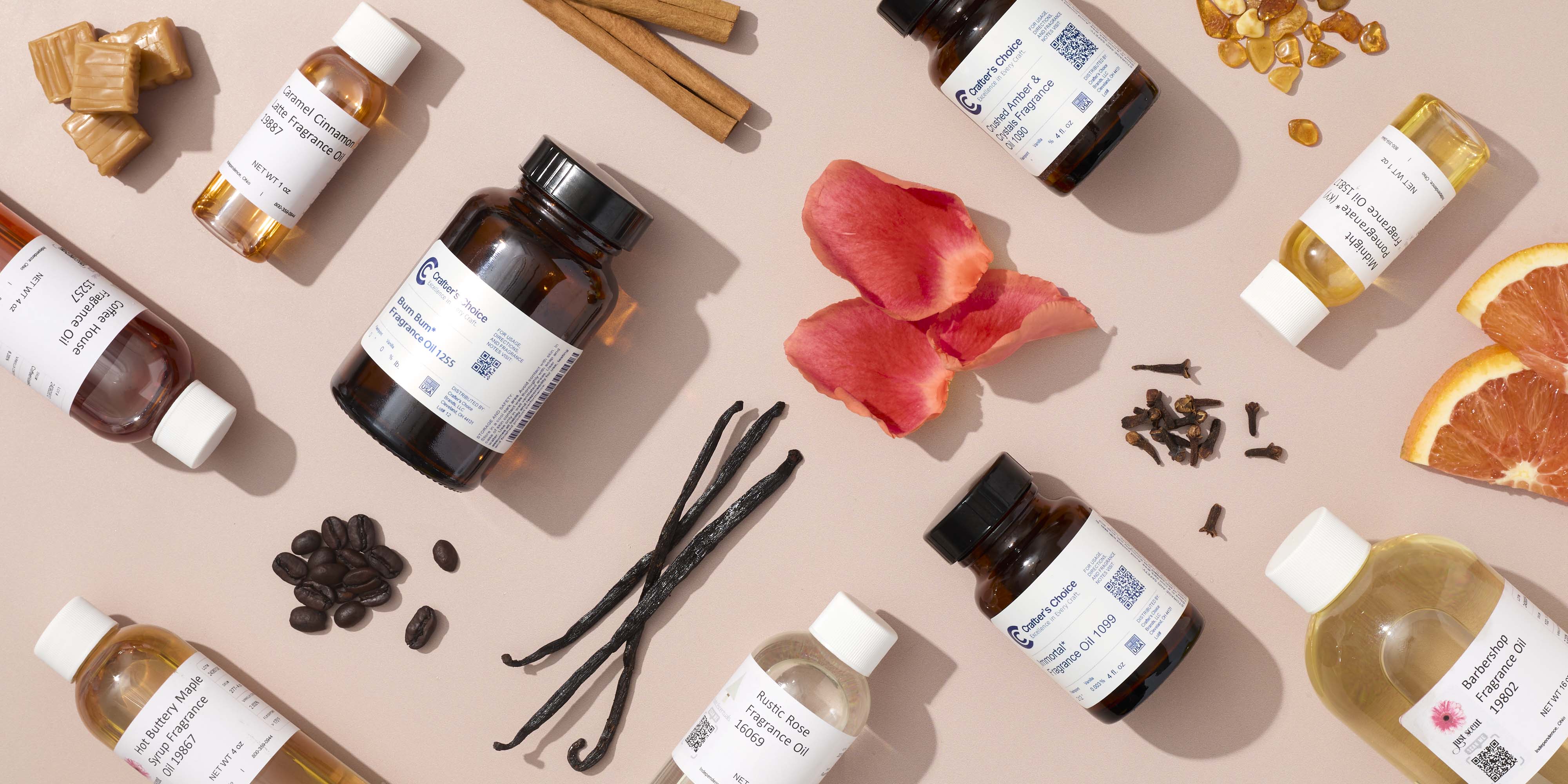 Flat lay of fragrance oil bottles arranged on a soft pink background with scent ingredients including cinnamon sticks, vanilla beans, coffee beans, rose petals, cloves, citrus slices, caramel pieces, and amber chips.