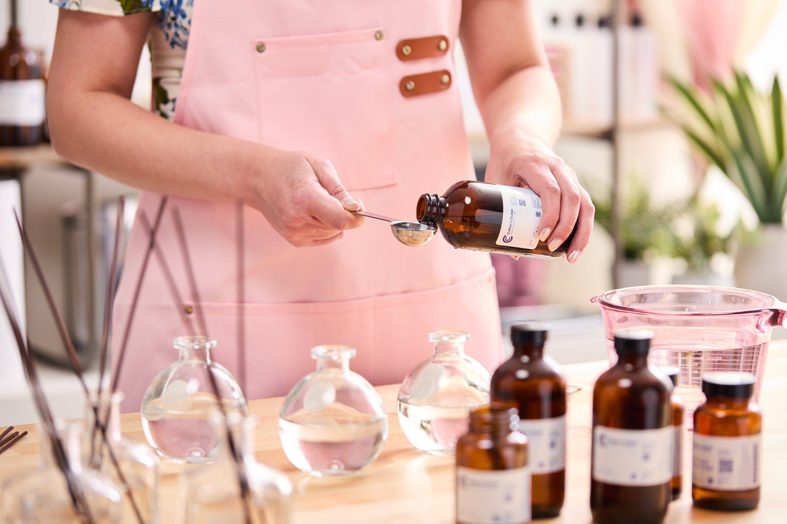 Person in a pink apron measuring fragrance oil from an amber bottle into a metal spoon, with round glass bottles filled with liquid and additional amber bottles arranged on a wooden workspace.