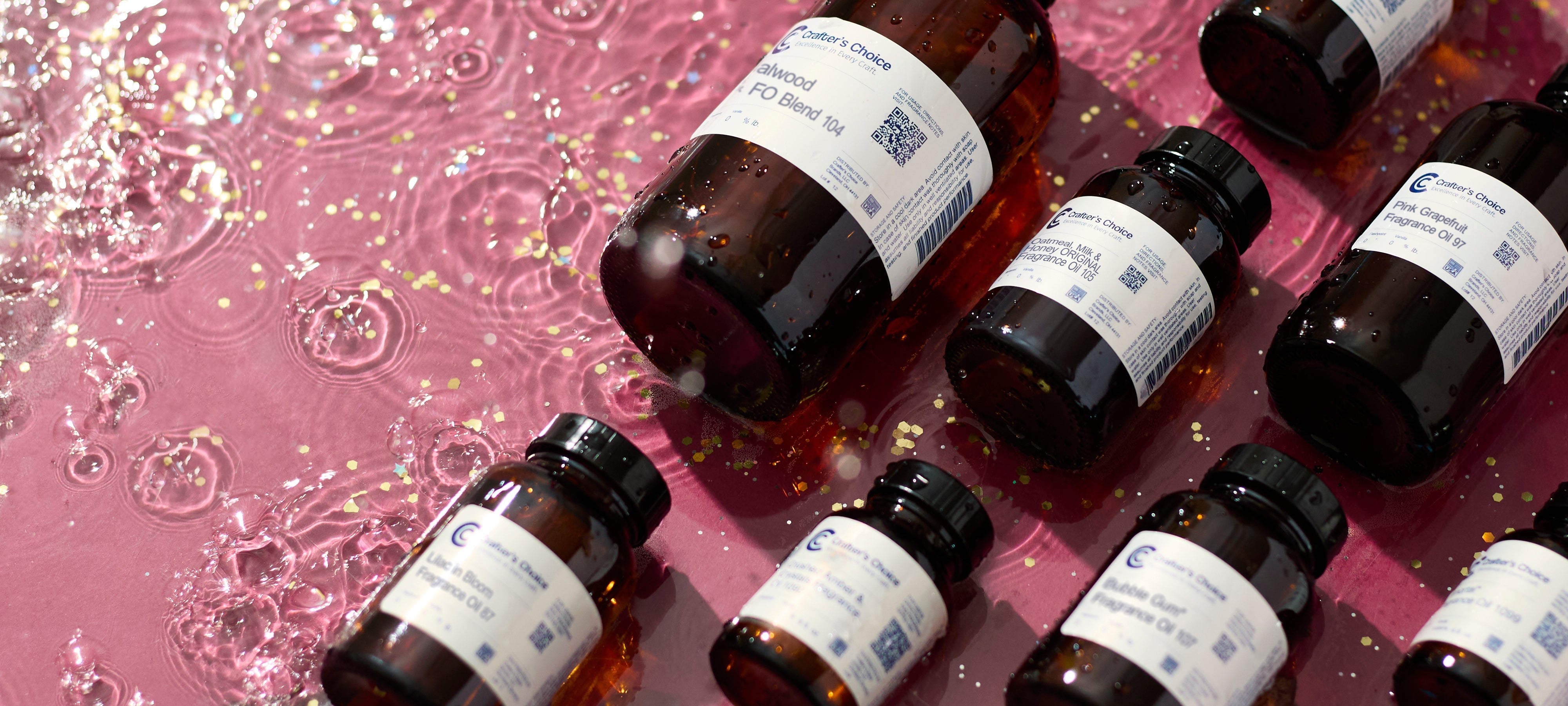 Amber Crafter’s Choice fragrance bottles arranged on a table beside clear diffuser bottles with reeds, while a person in a pink apron measures fragrance oil in the background