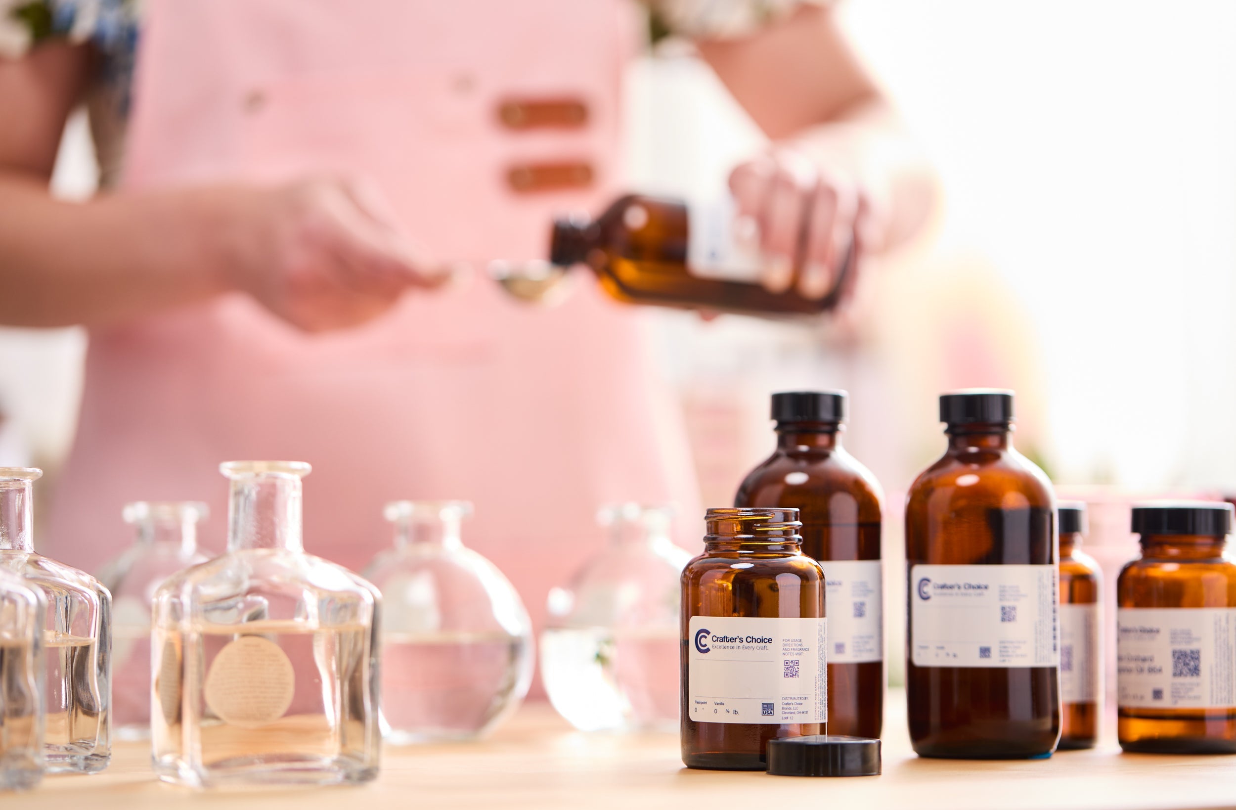 Amber glass fragrance oil bottles and clear containers filled with liquid sit on a worktable while a person in a pink apron measures fragrance oil with a spoon in a bright crafting studio.