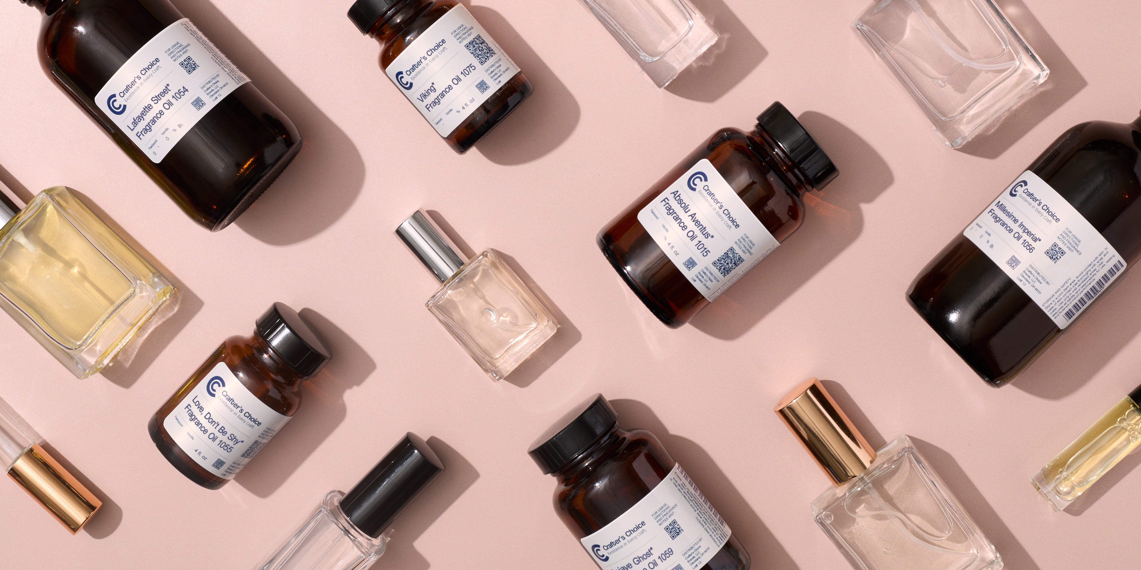 Flat lay of fragrance oils and perfume bottles on a soft pink background, featuring amber glass bottles with labeled scents alongside clear glass spray bottles filled with light-colored fragrance.