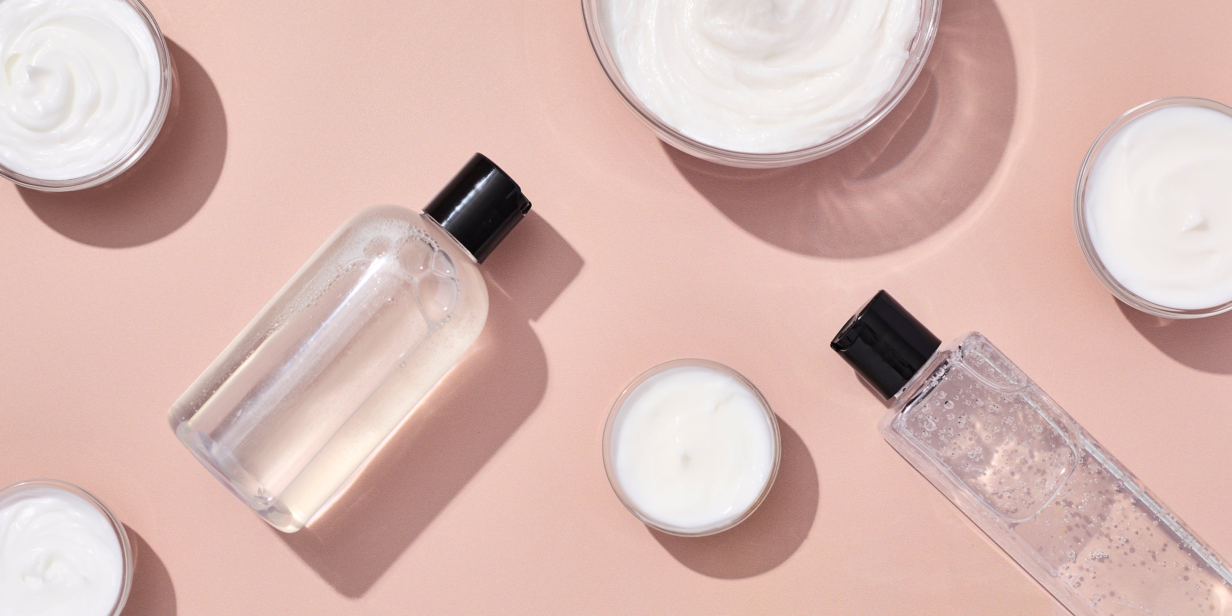 Flat lay of skincare bases including clear bottles of liquid gel and small bowls of white cream arranged on a soft peach background.