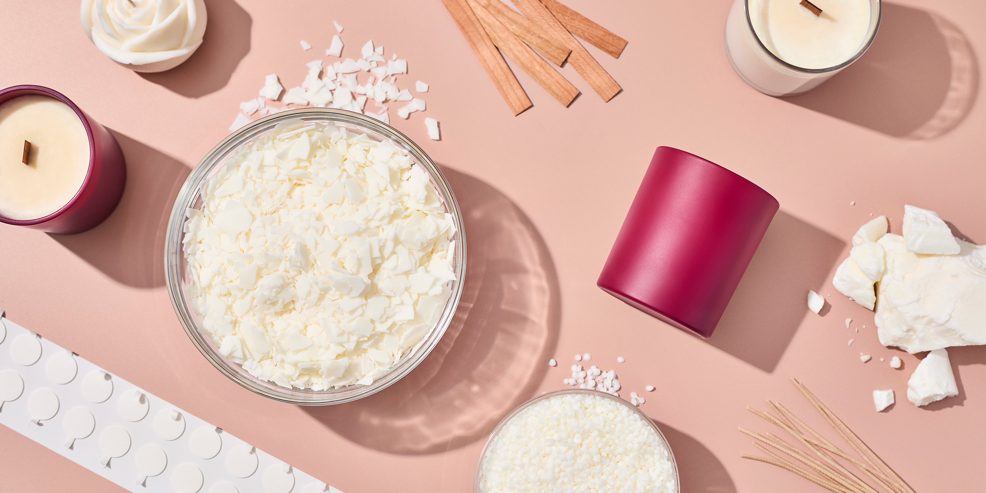 Flat lay of candle-making supplies on a blush surface, including soy wax flakes, wax chunks, wick stickers, wooden wicks, wicks, a berry-colored candle jar, finished candles, and a bowl of wax pellets.