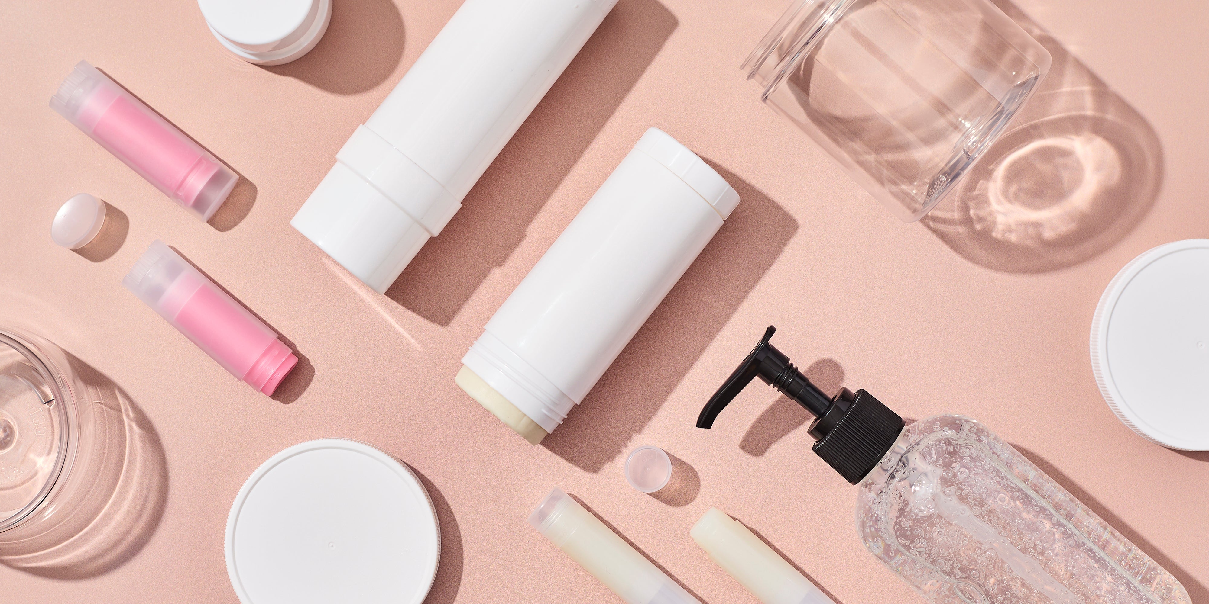 Flat lay of empty cosmetic packaging including white deodorant tubes, clear lip balm tubes, a clear pump bottle, plastic jars, and lids arranged on a soft peach background.