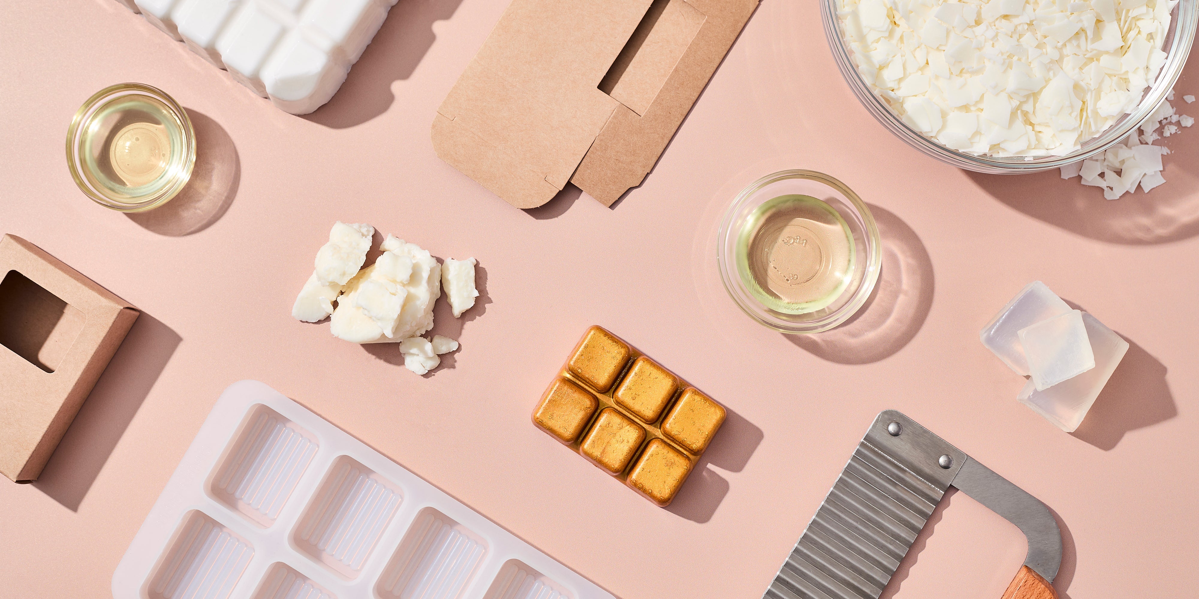 Flat lay of soap-making supplies on a blush surface, including melt-and-pour soap bases, fragrance oil in glass bowls, kraft paper boxes, silicone molds, a wavy soap cutter, and finished gold soap cubes.