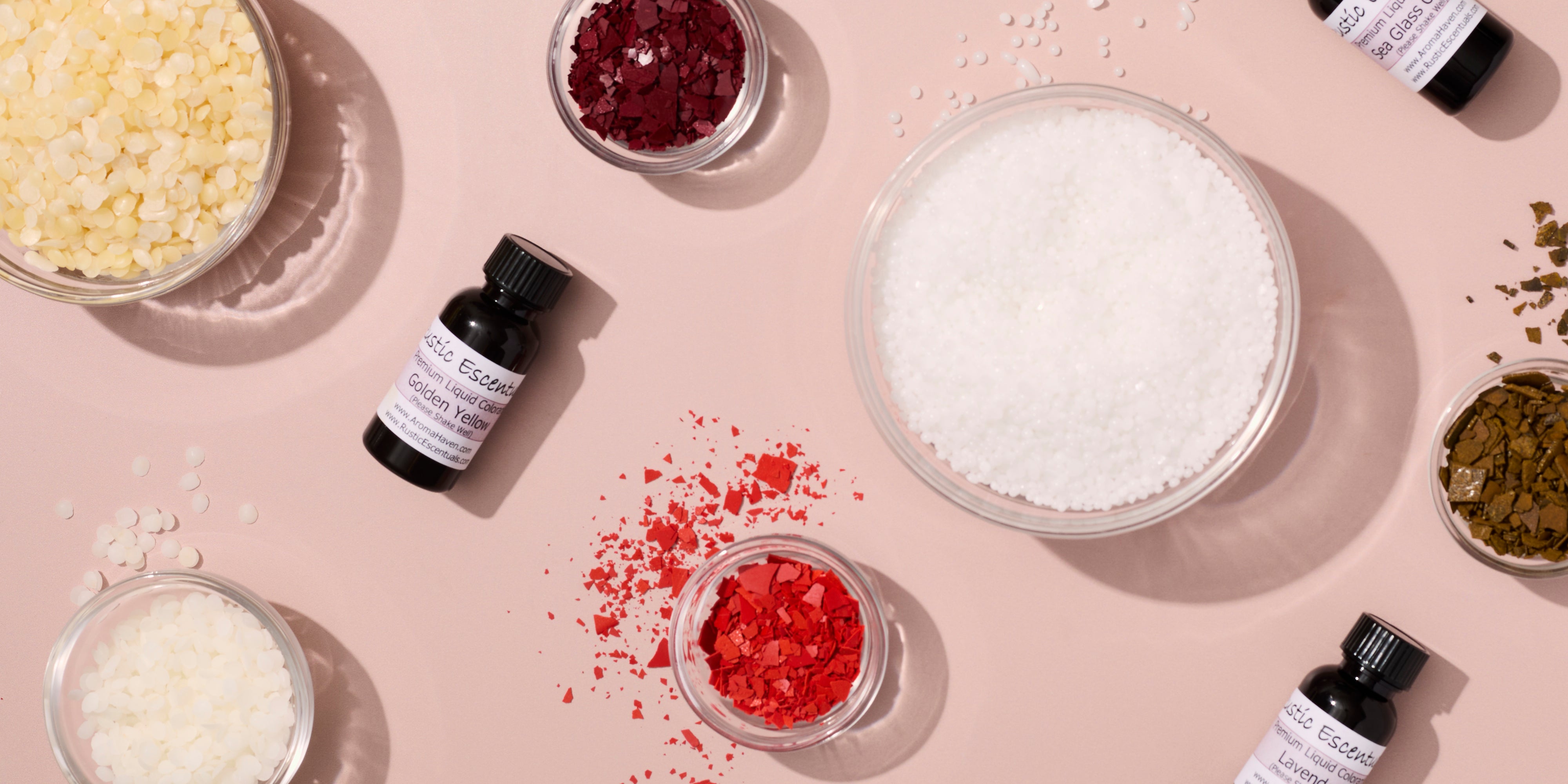 Flat lay of candle making additives and colorants on a soft pink background, including bowls of wax flakes, granules, and pigment chips in white, red, yellow, and green, alongside small bottles of liquid candle dye.