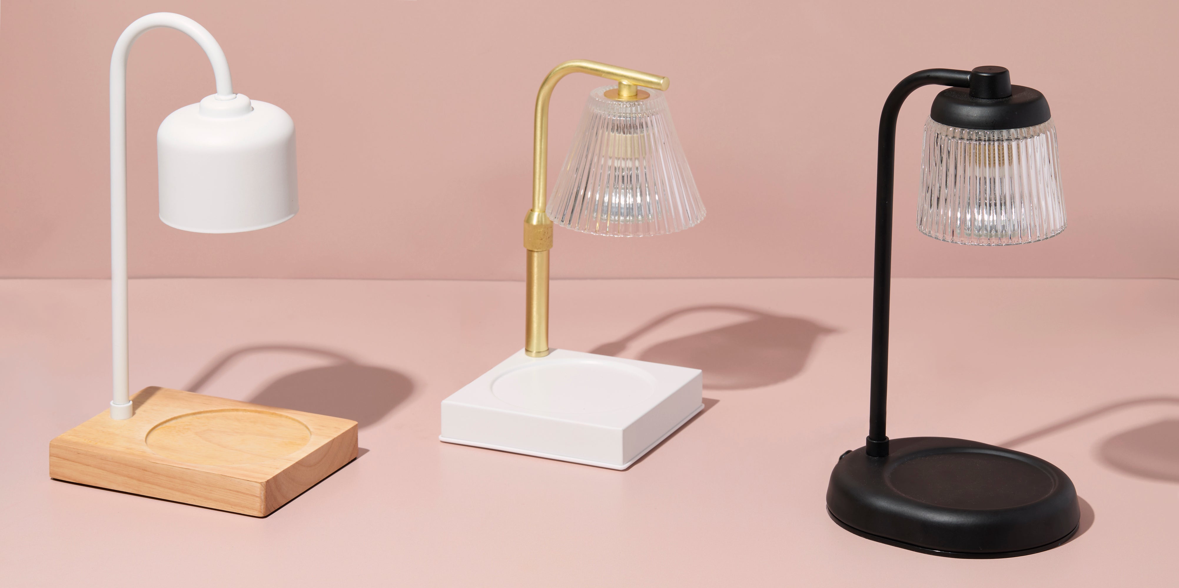 Three candle warmers displayed on a soft pink background, featuring modern designs in white with a wood base, brass with a white base, and black with a matching base, each with a curved arm and glass shade.