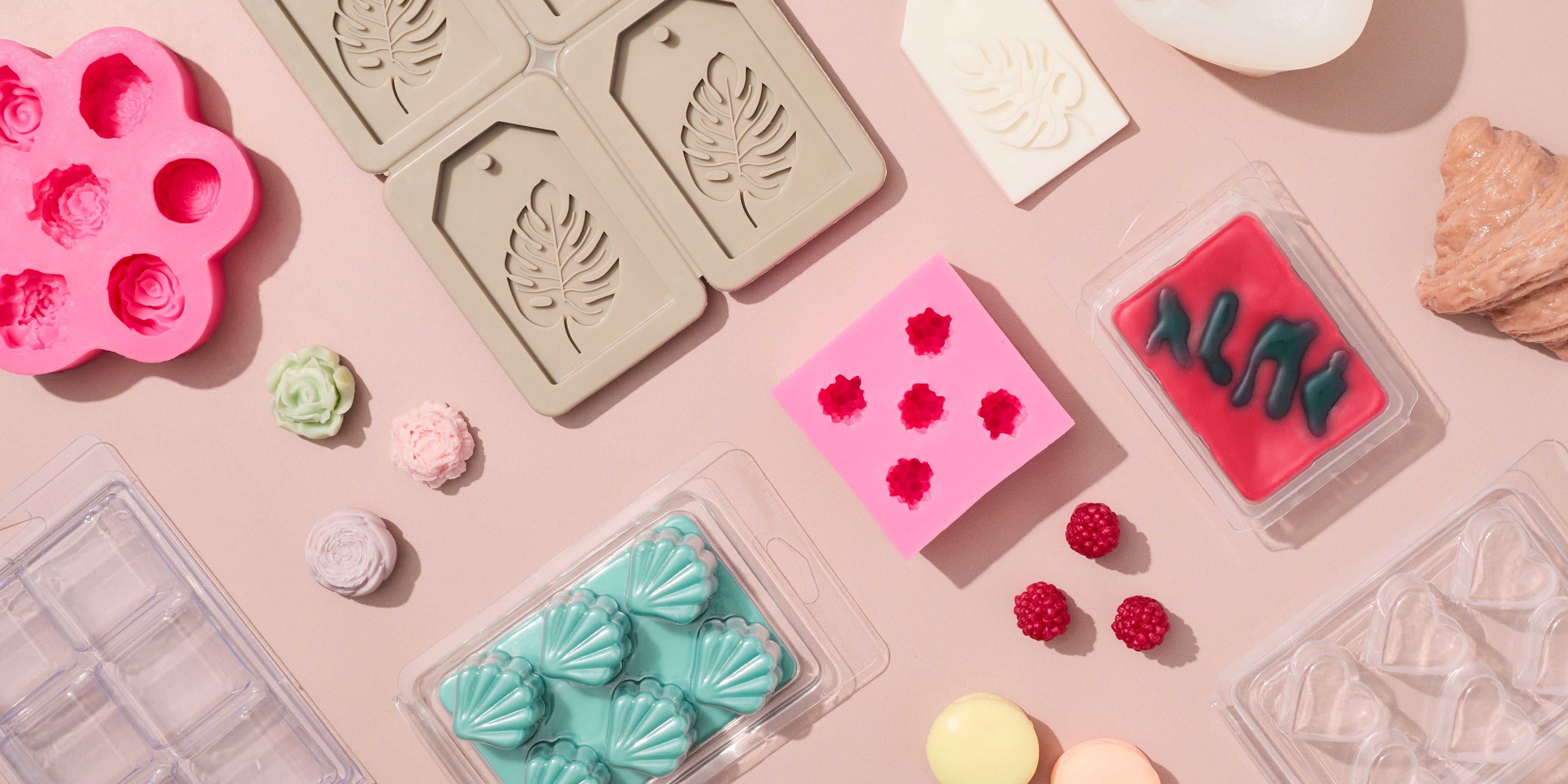 Flat lay of soap and wax melt molds and clamshell packaging on a soft pink background, featuring floral, leaf, shell, heart, and geometric shapes in pastel colors, with finished melts displayed alongside.