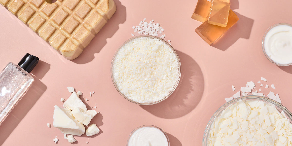 Flat lay of personal care bases including white wax flakes, pellets, and chunks, clear gel in a bottle, amber soap cubes, a waffle soap bar, and bowls of cream arranged on a soft peach background.