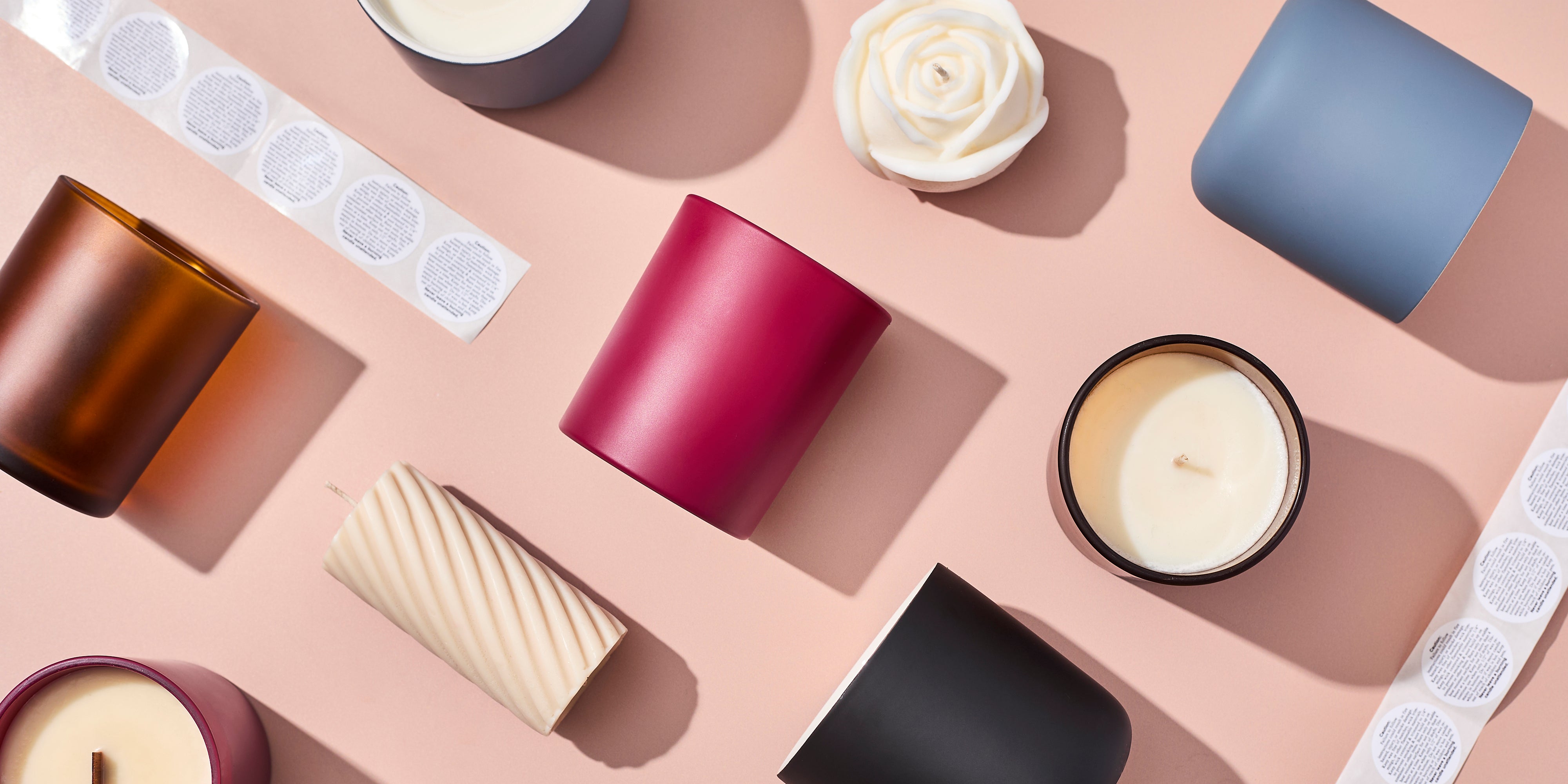 Flat lay of assorted candle jars and finished candles on a blush surface, including matte glass vessels in various colors, a rose-shaped candle, a pillar candle, and safety label stickers.