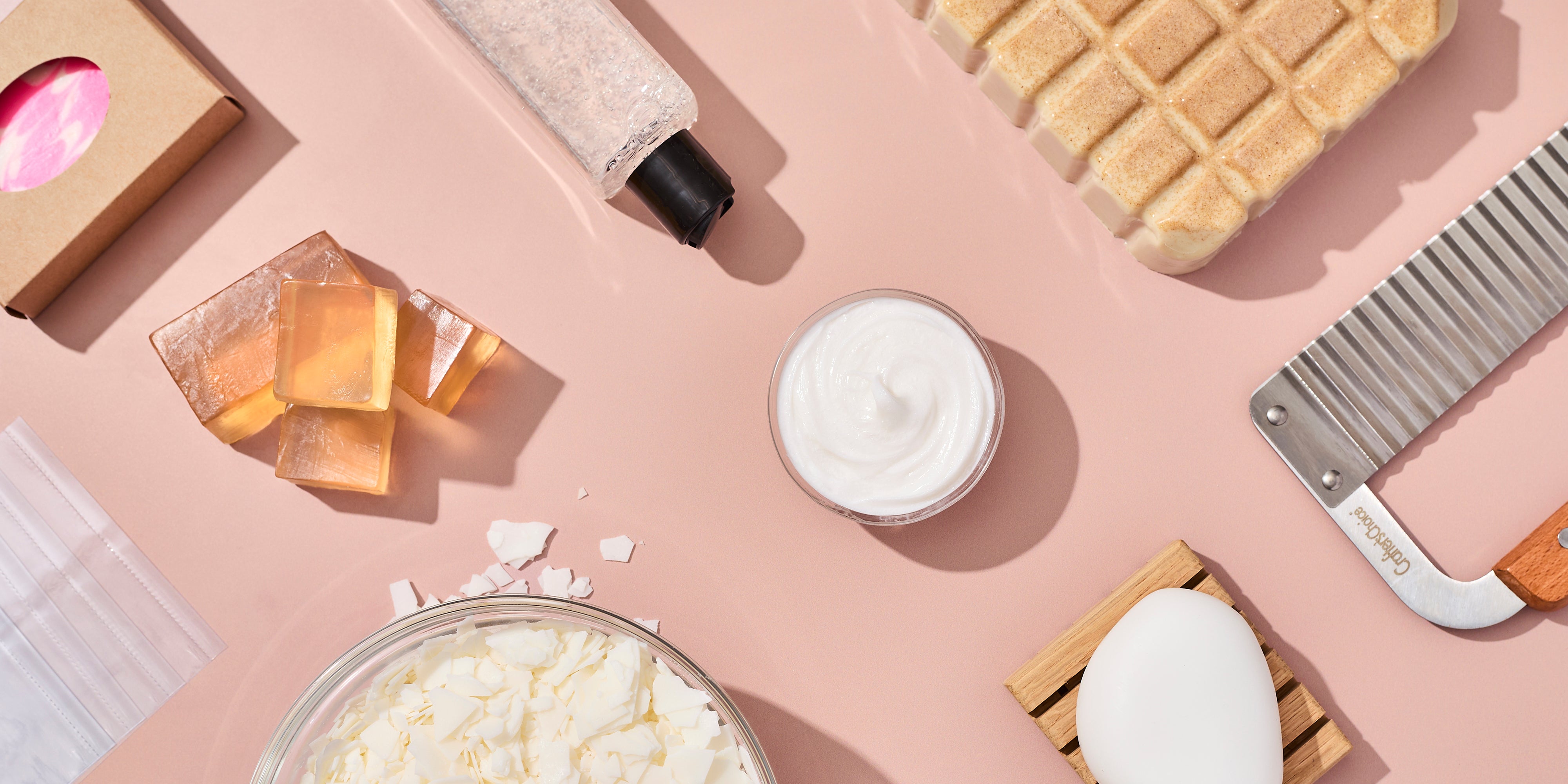 Flat lay of soap-making supplies on a blush surface, including clear soap cubes, a swirl bar in kraft packaging, exfoliating gel in a tube, whipped body cream, soap flakes in a bowl, a wooden soap dish, a finished bar, and a wavy soap cutter.