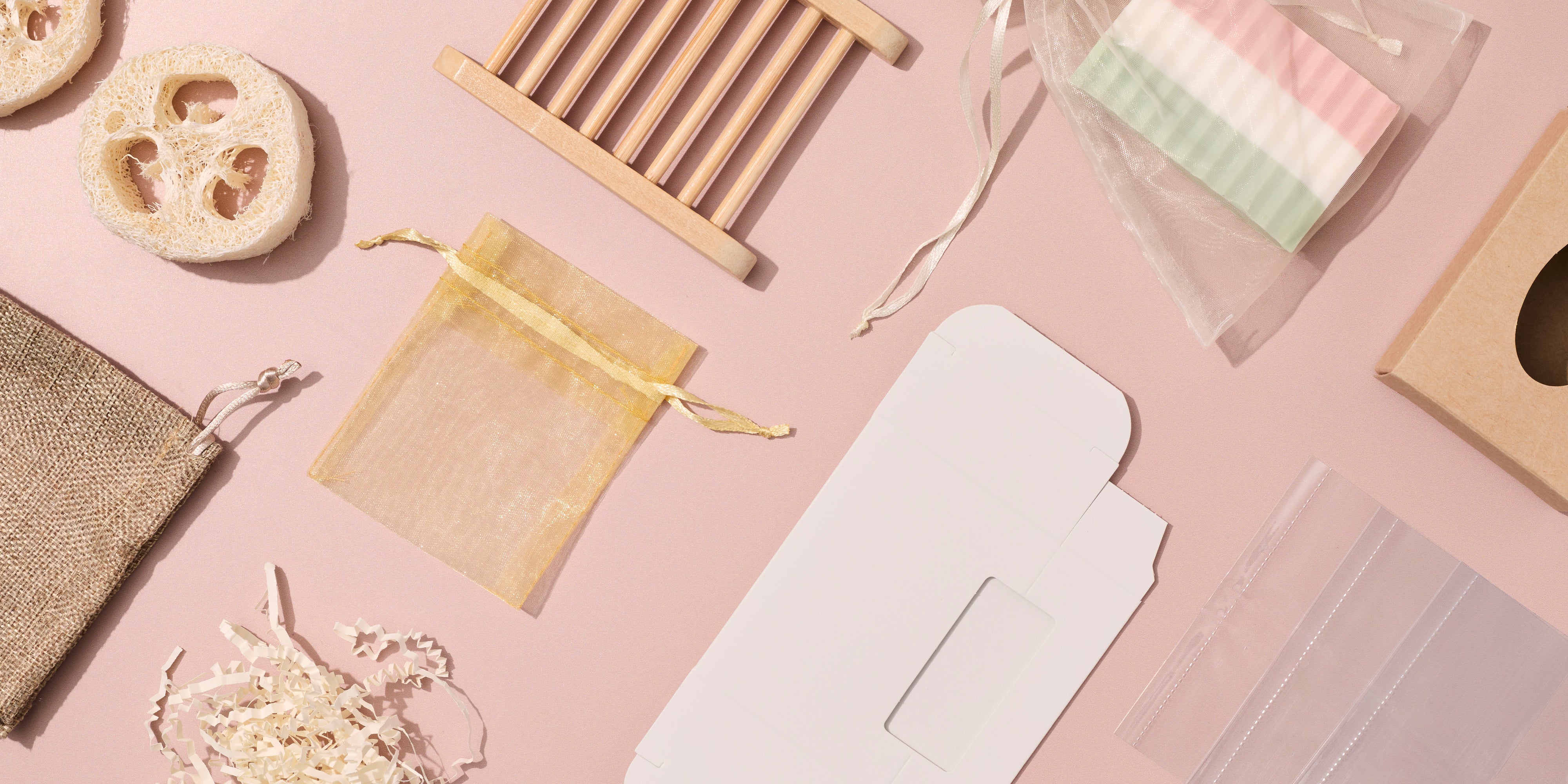 Flat lay of soap and candle packaging accessories on a soft pink background, including organza and burlap bags, gift boxes, soap cutters, filler paper, and natural exfoliating sponges arranged neatly.