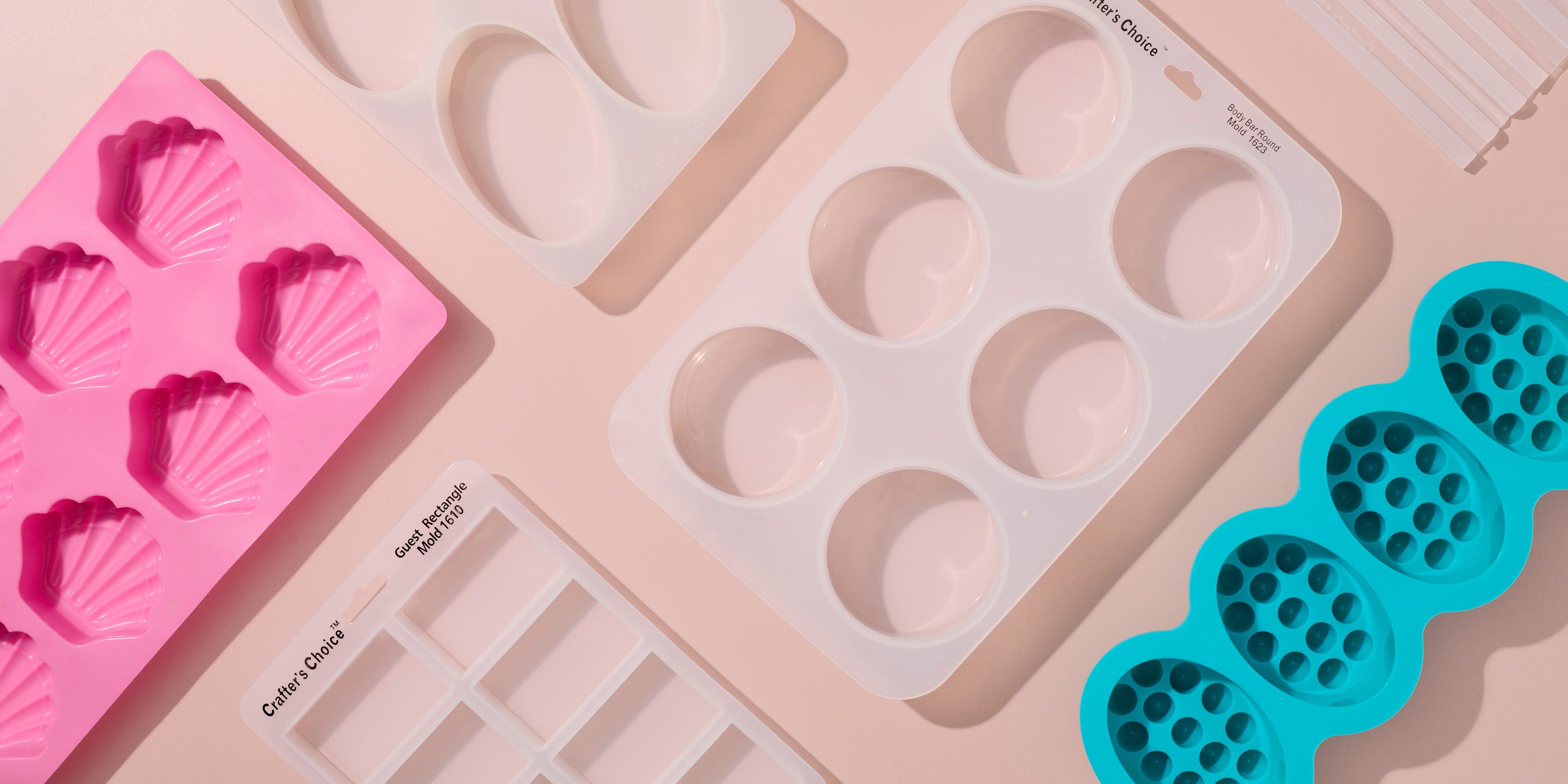 Flat lay of colorful silicone soap molds on a soft neutral background, featuring shell shapes, hearts, rounds, rectangles, and textured designs in pink, white, and teal.