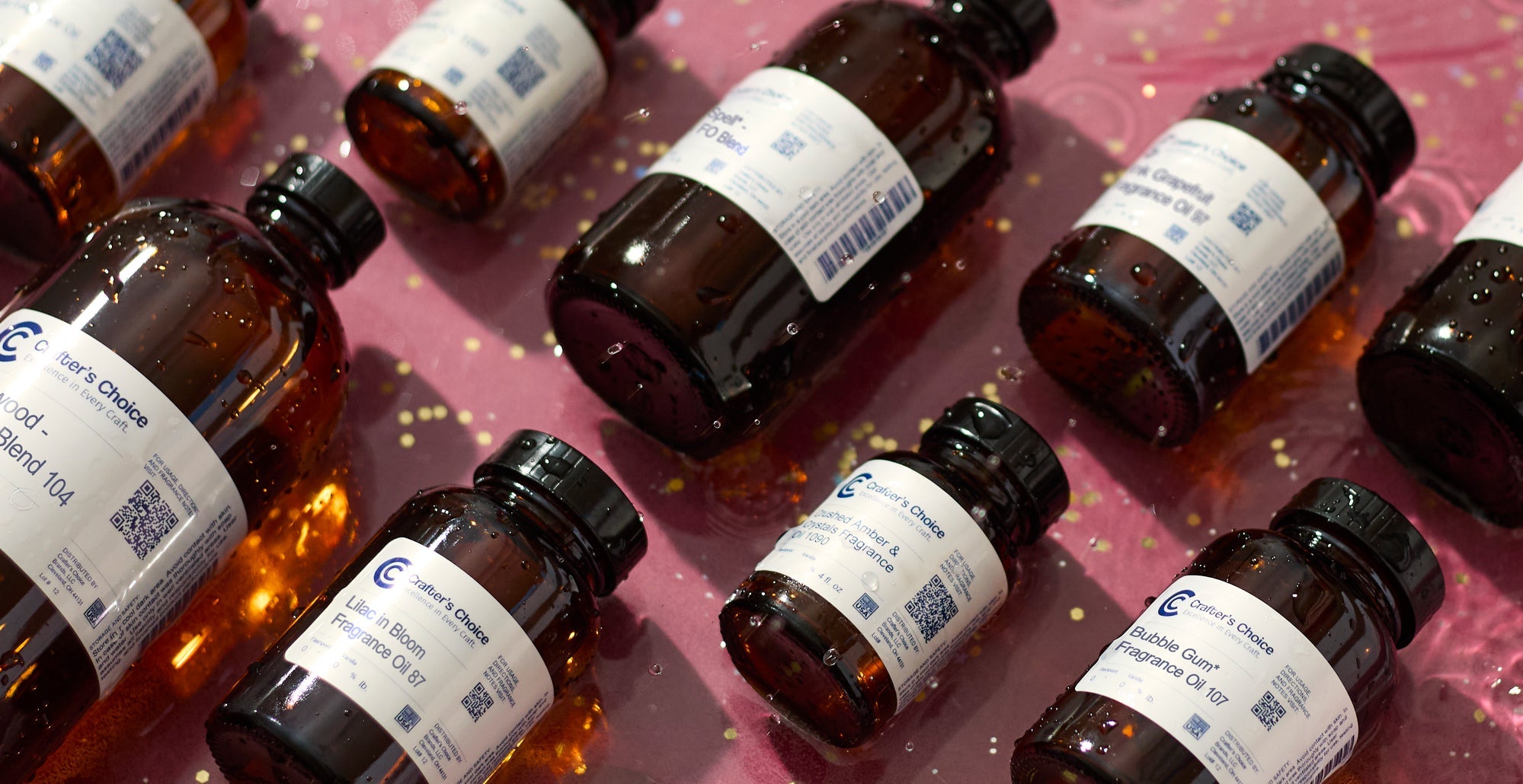 Assorted Crafter’s Choice fragrance oil bottles scattered on a pink surface with water droplets and reflective highlights.