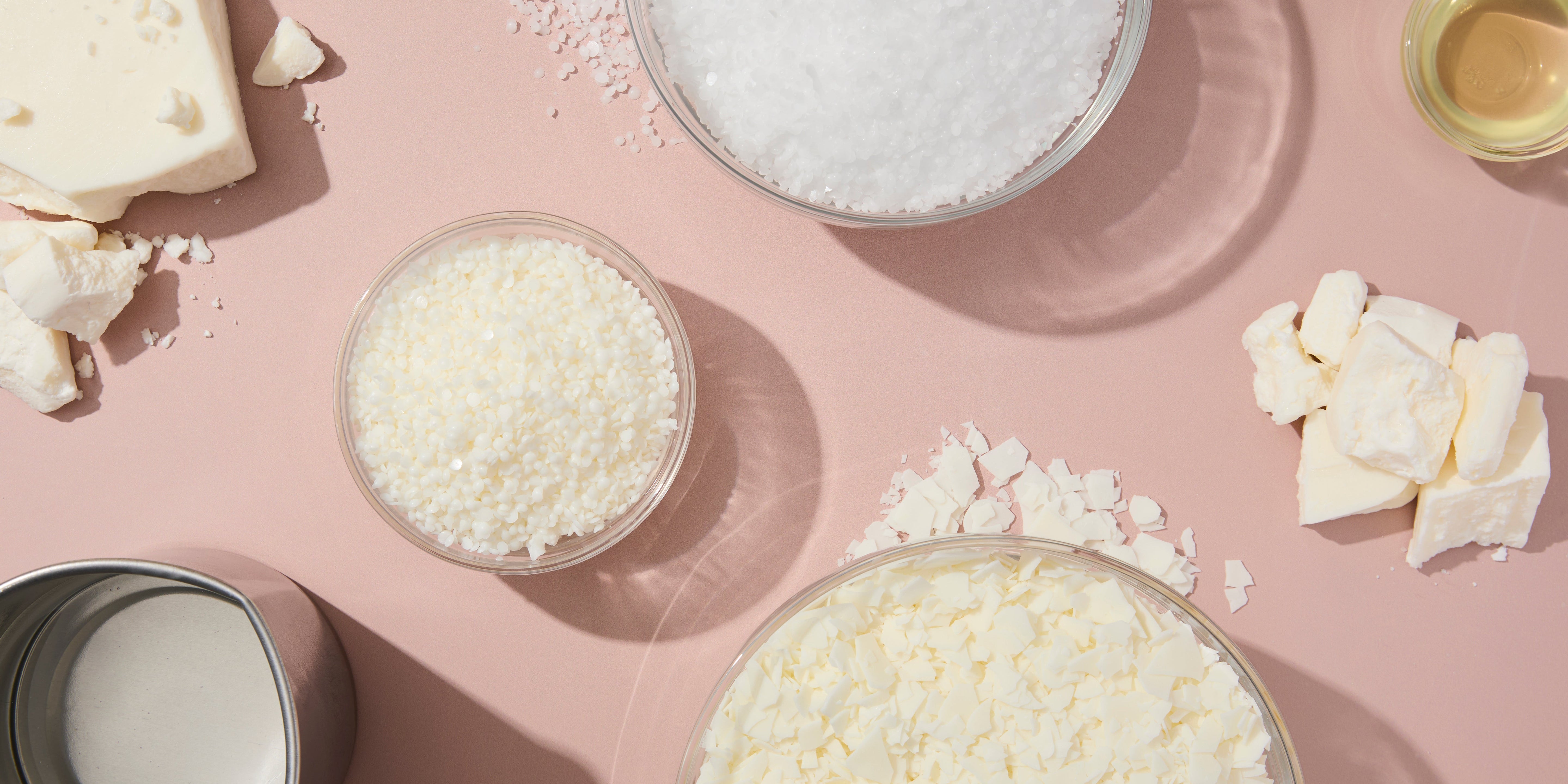 Flat lay of various candle wax types on a blush surface, including soy flakes, wax pellets, wax chunks, and melted wax in a tin, arranged in clear bowls and scattered around the workspace.