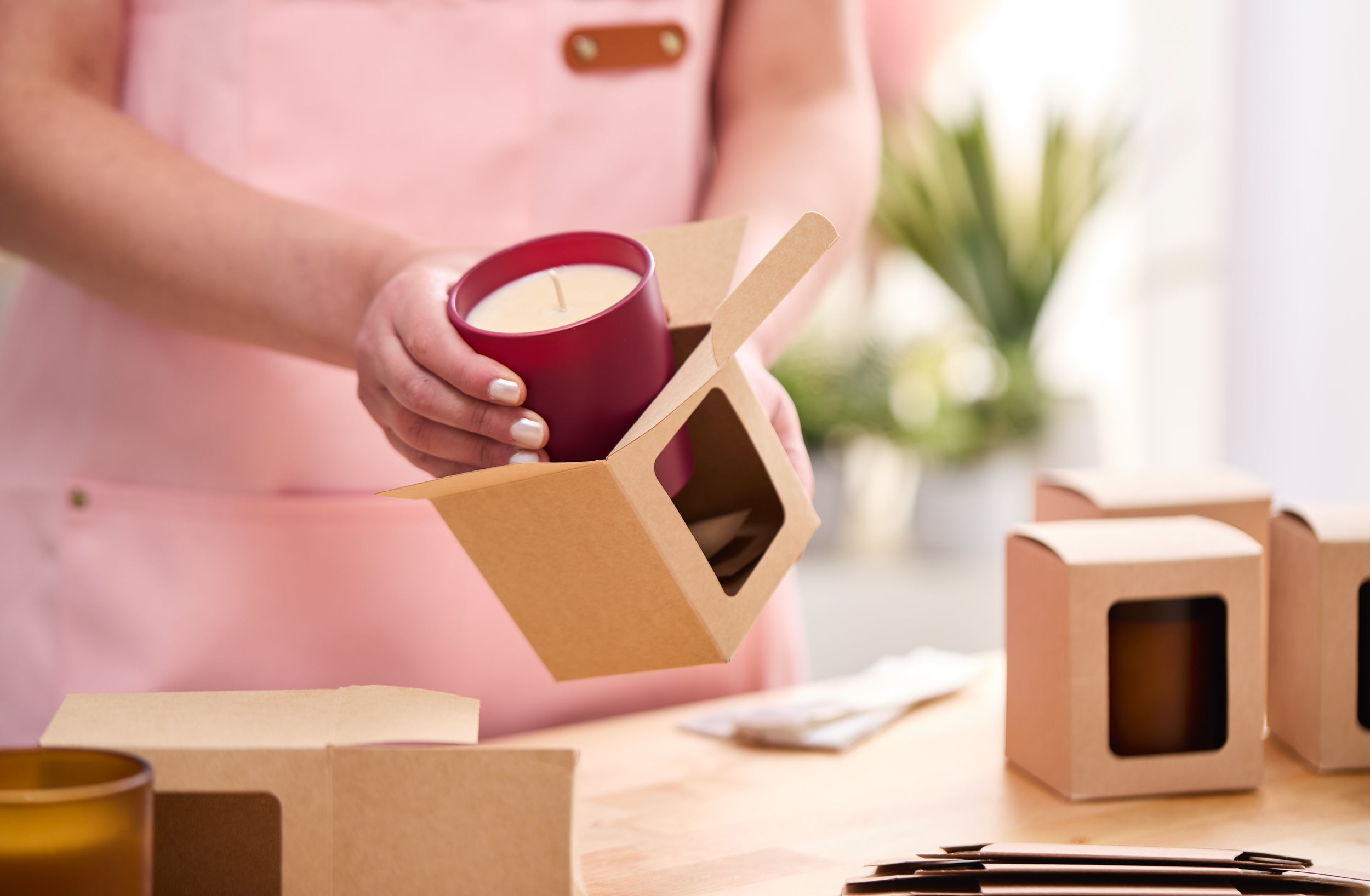 Person in a pink apron placing a finished candle in a burgundy jar into a kraft paper display box on a wooden table, with other assembled and flat-packed boxes nearby in a bright workspace.