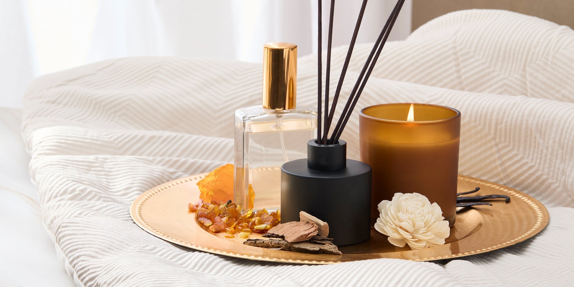 Gold tray on a bed holds a glass perfume bottle, black reed diffuser, lit amber candle, crystals, wood pieces, and a carved flower in a calm, spa-like setting.