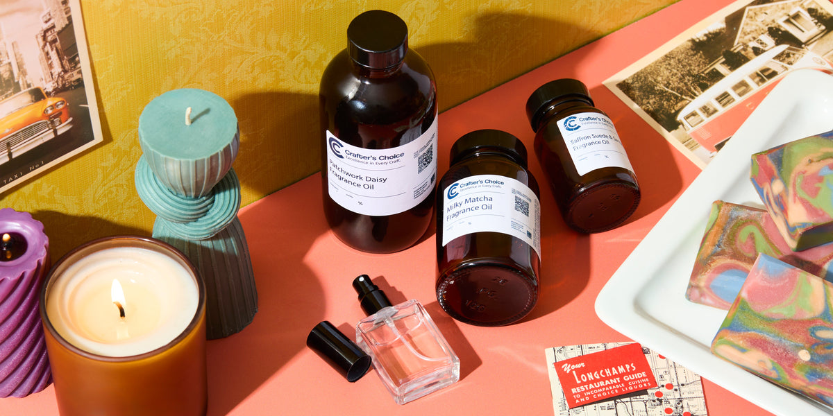 Lit candle, sculptural blue candle, fragrance oil bottles, perfume bottle, and colorful handmade soap bars arranged on a tabletop with postcards, creating a bright, artisanal candle- and soap-making scene.