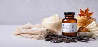 Amber bottle of Cloud Dancer fragrance oil displayed with vanilla beans, whipped cream, caramel, and whipped topping on a soft cream-colored cloth, accented by an autumn leaf.