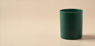 Dark green cylindrical candle vessel on a warm beige background with soft lighting and a subtle shadow.
