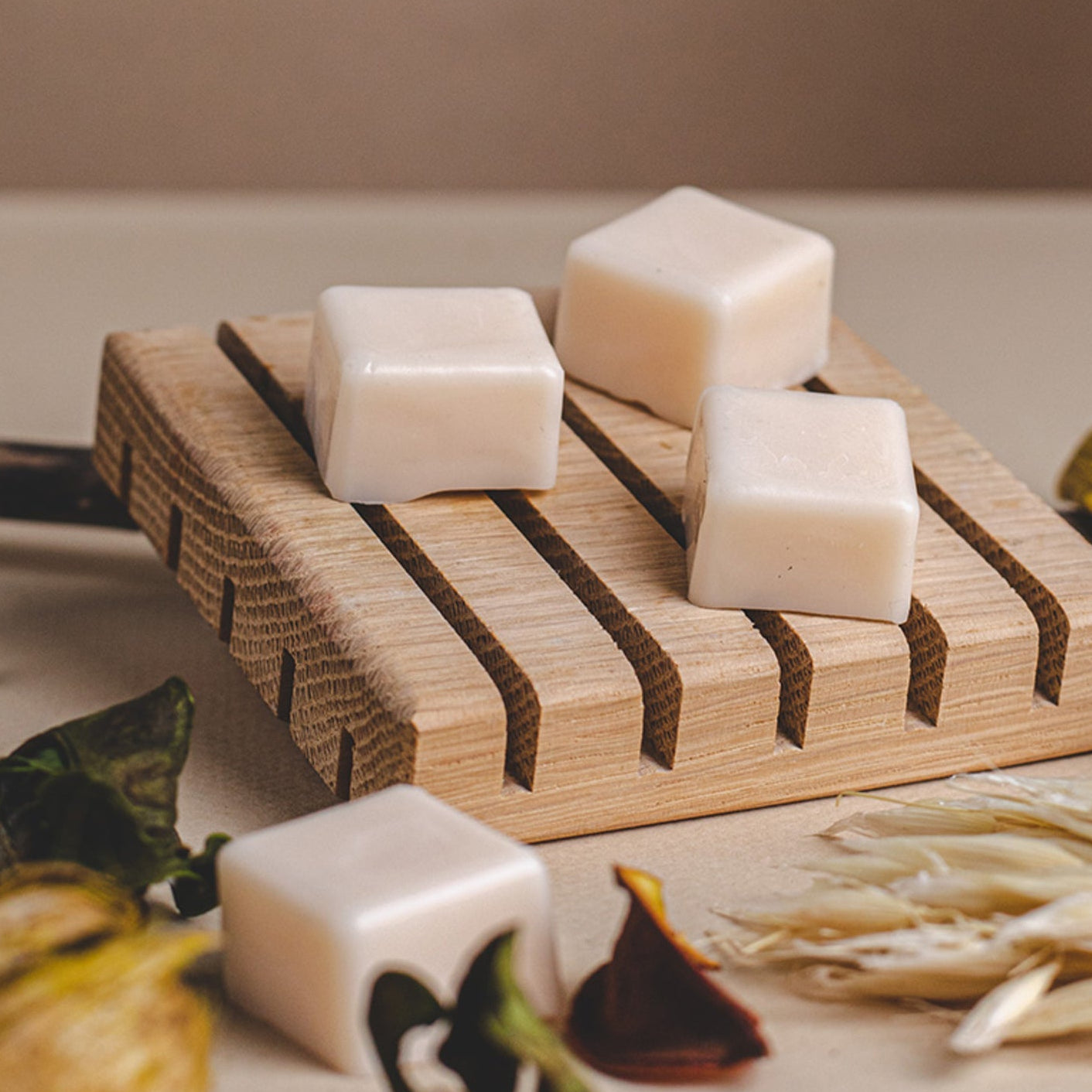 White cube-shaped wax melts arranged on a wooden tray, with one melt placed nearby. Dried botanical elements and seed pods surround the tray on a neutral surface, creating a warm, natural home fragrance scene.