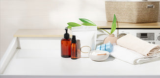 Amber pump and spray bottles, a white pouch, blue cubes, and a bowl of powder beside folded towels on a laundry room counter.