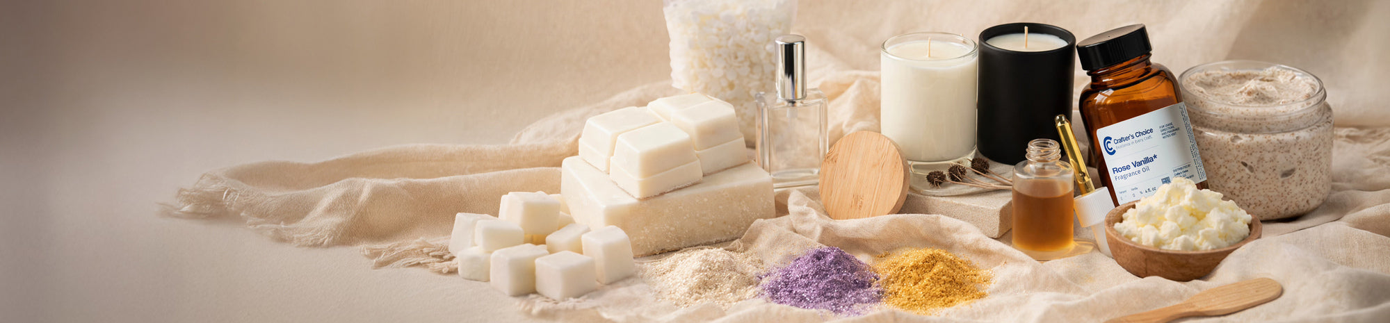 Neutral-toned display of soap making supplies including melt-and-pour soap cubes, fragrance oils, candles, shea butter, botanical powders, and a jar of scrub arranged on soft fabric for a clean, natural aesthetic.