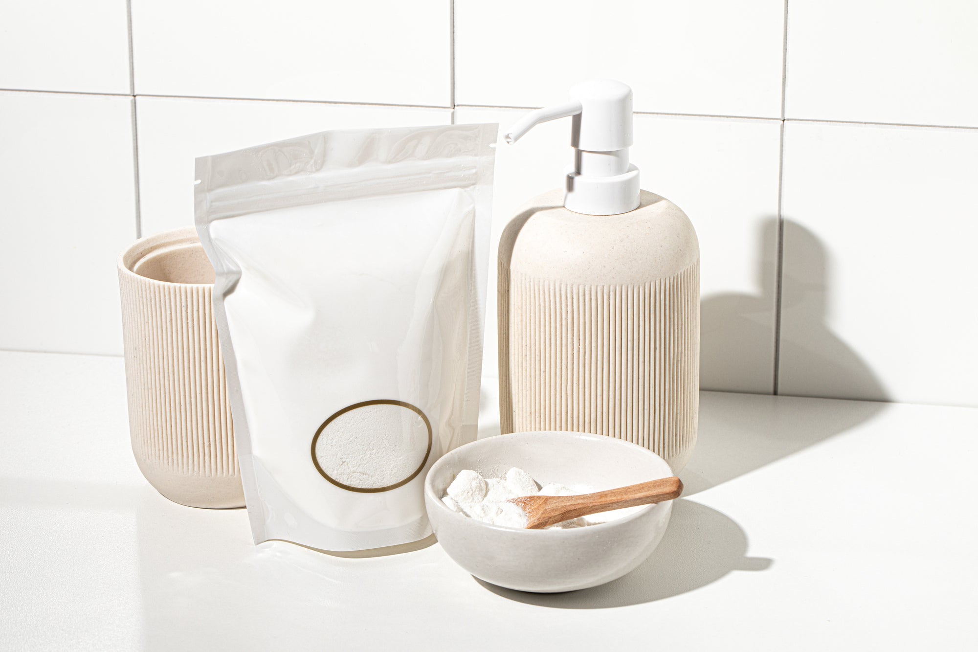 Minimalist laundry care products displayed on a white surface, including a refill pouch of powdered laundry detergent, a ceramic soap dispenser, a matching storage vessel, and a small bowl of detergent with a wooden scoop, set against a tiled background.