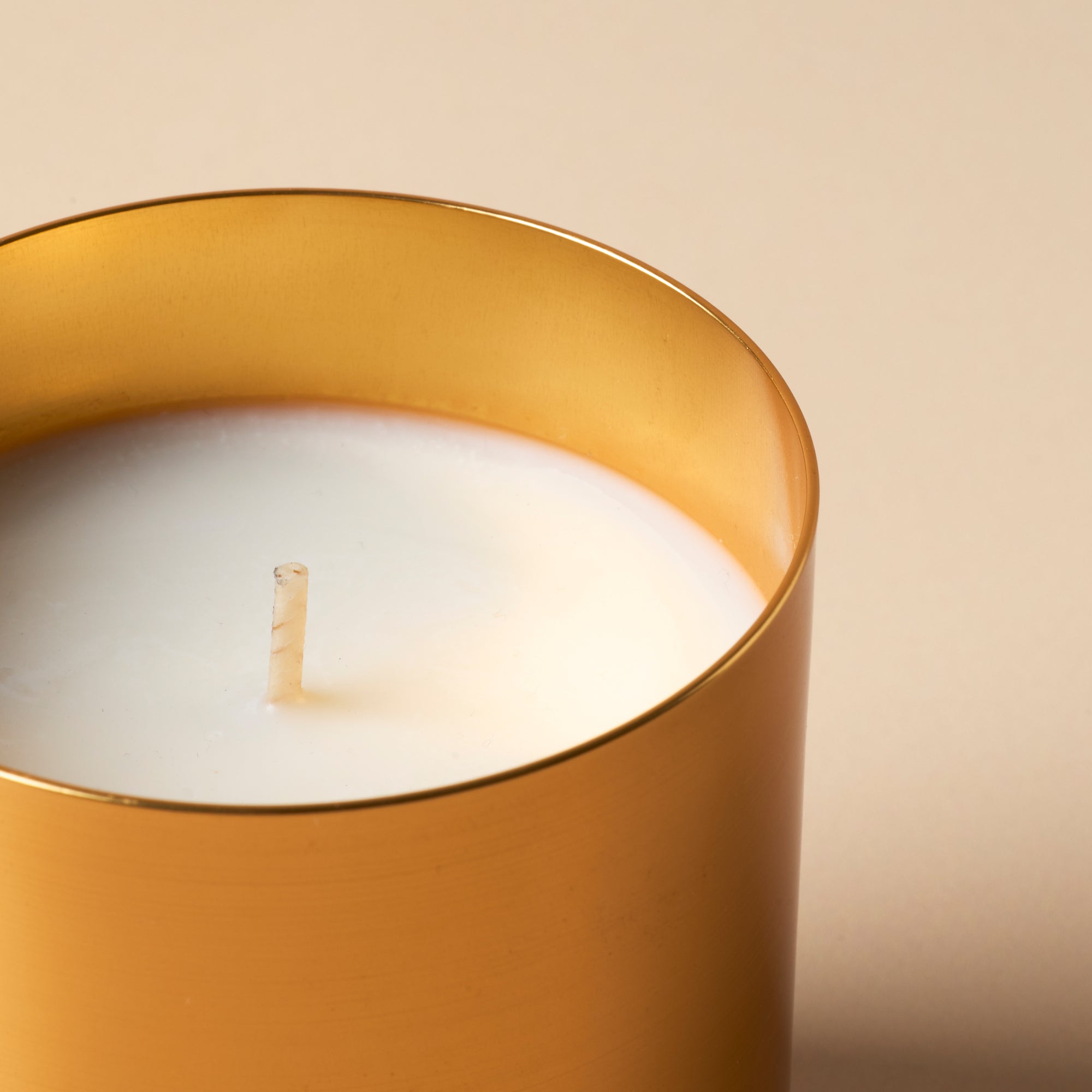 candle sits against a softly lit neutral background, highlighting the clean wax surface and warm metallic finish.