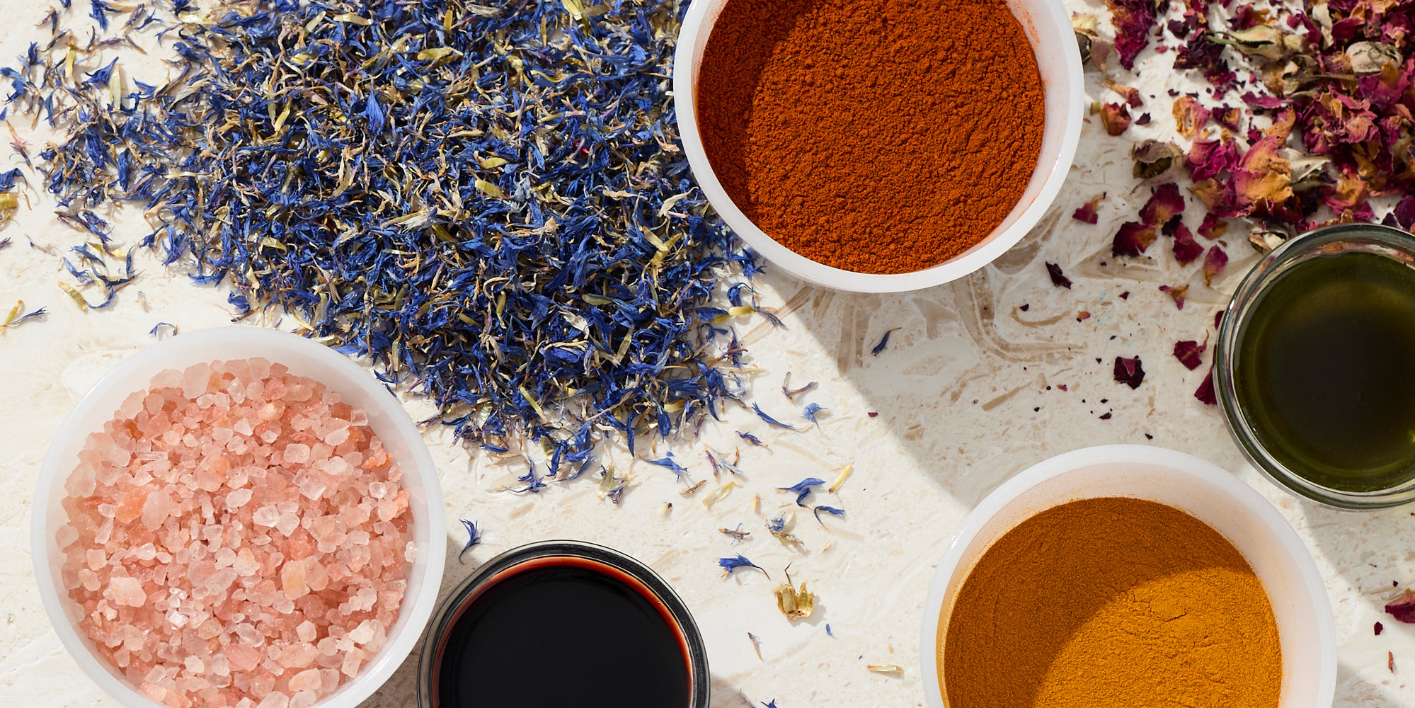 Flat lay of natural additives and botanicals including dried lavender and blue petals, pink Himalayan salt, powdered clays, spices, and small bowls of oils arranged on a light textured surface.